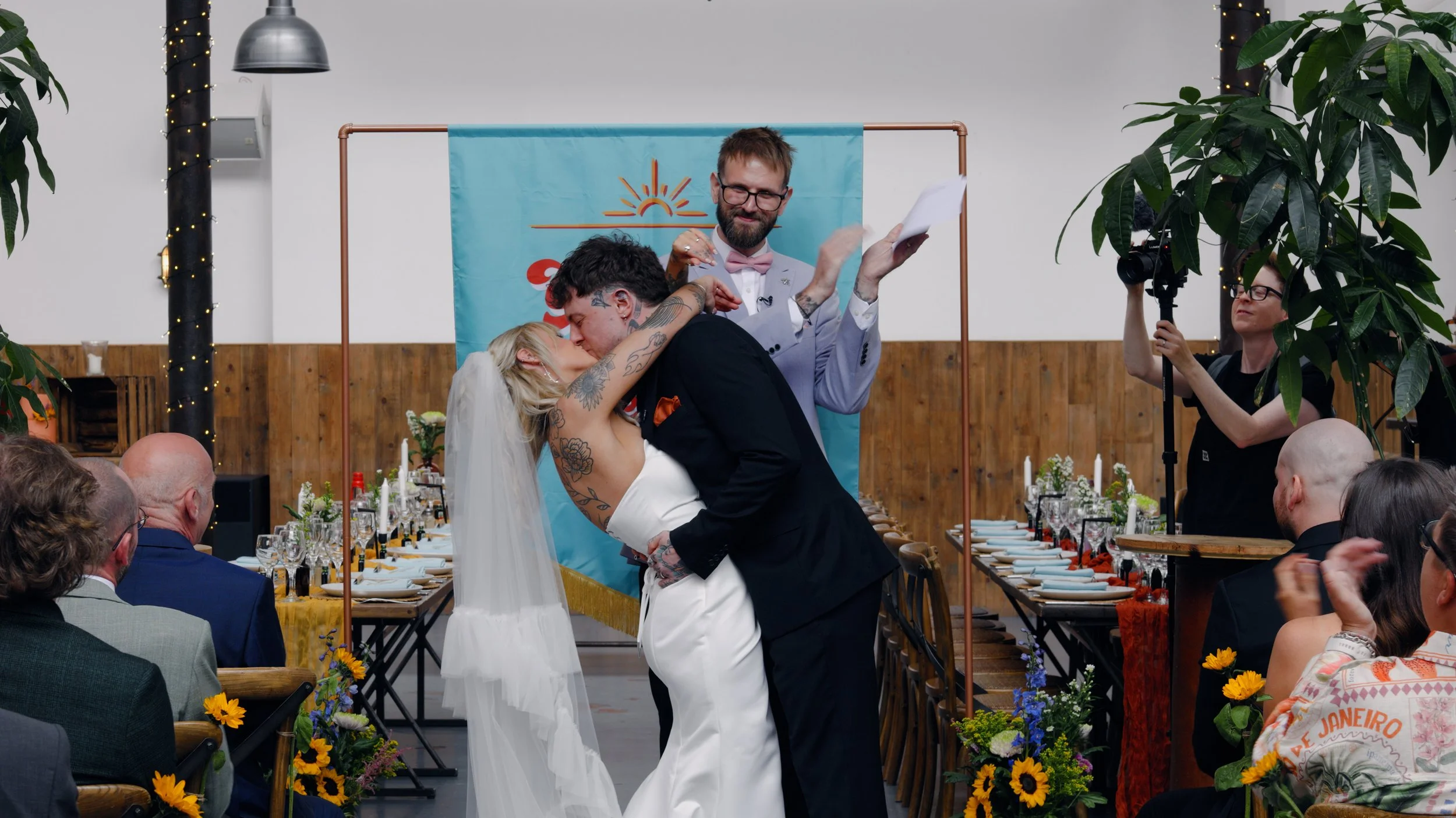 The groom in an all in black wedding suit kisses the bride in a colourful wedding at the River Mill in Derby, taken by wedding videographer Laura Baker Productions