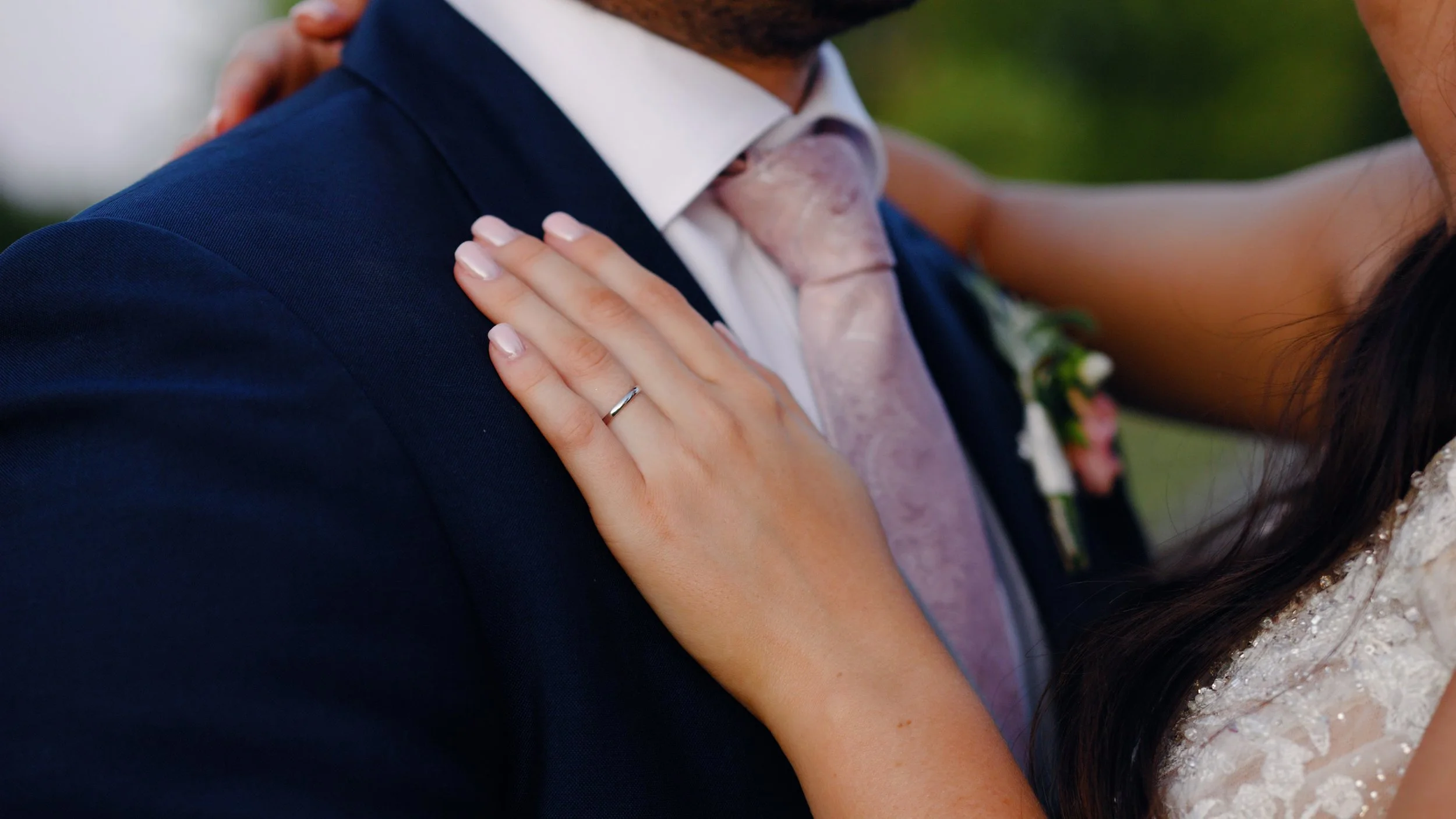 The bride shows off her wedding ring hand against the groom's navy suit and pink tie combination, taken by wedding videographer laura baker productions