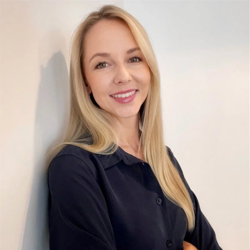 A young woman with long blonde hair, fair skin, and green eyes smiling, wearing a black shirt, standing against a plain light-colored wall.