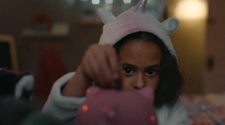 Child in unicorn hoodie holding a pink piggy bank.