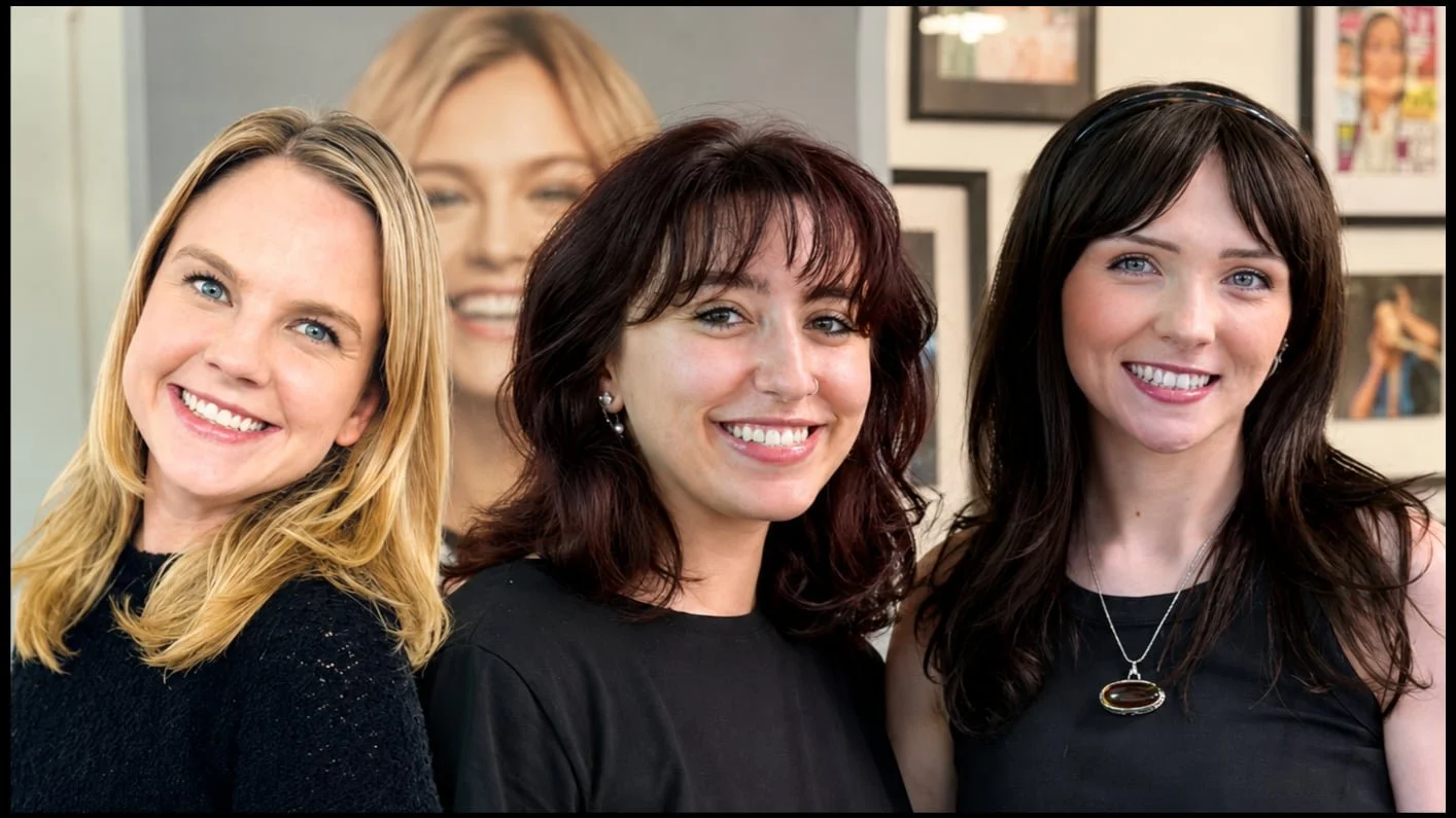Three smiling women pose together indoors with framed photos and a large portrait in the background. These are the bLUXE apprentice team offering specially reduced pricing for luxury services