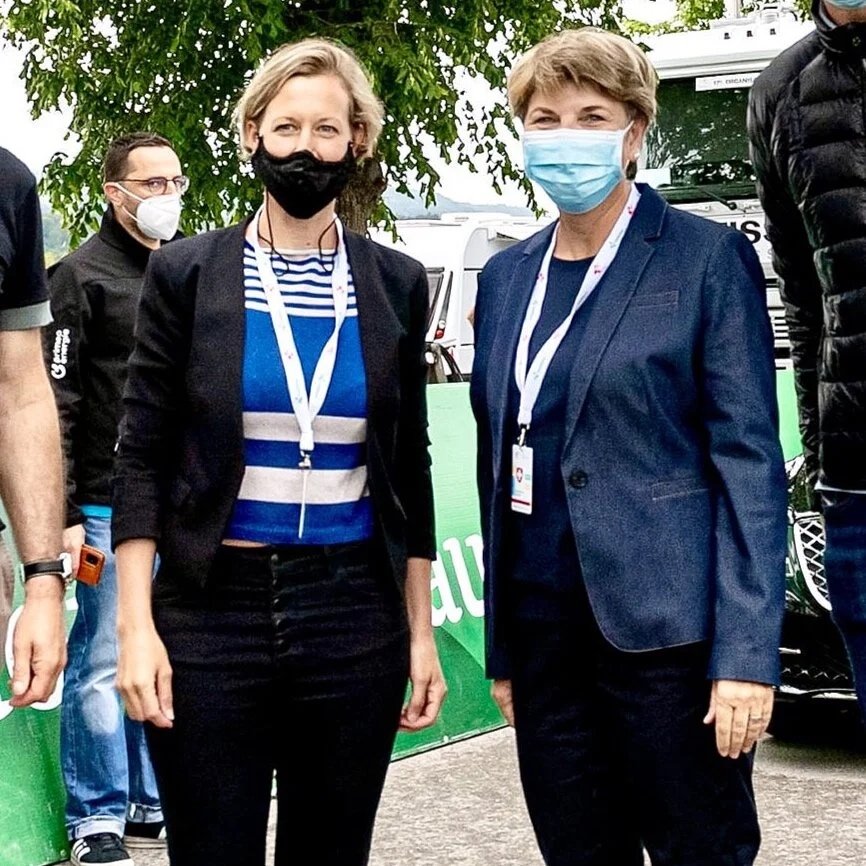 Zwei Frauen in formeller Kleidung tragen Masken und stehen draußen vor einigen Fahrzeugen.