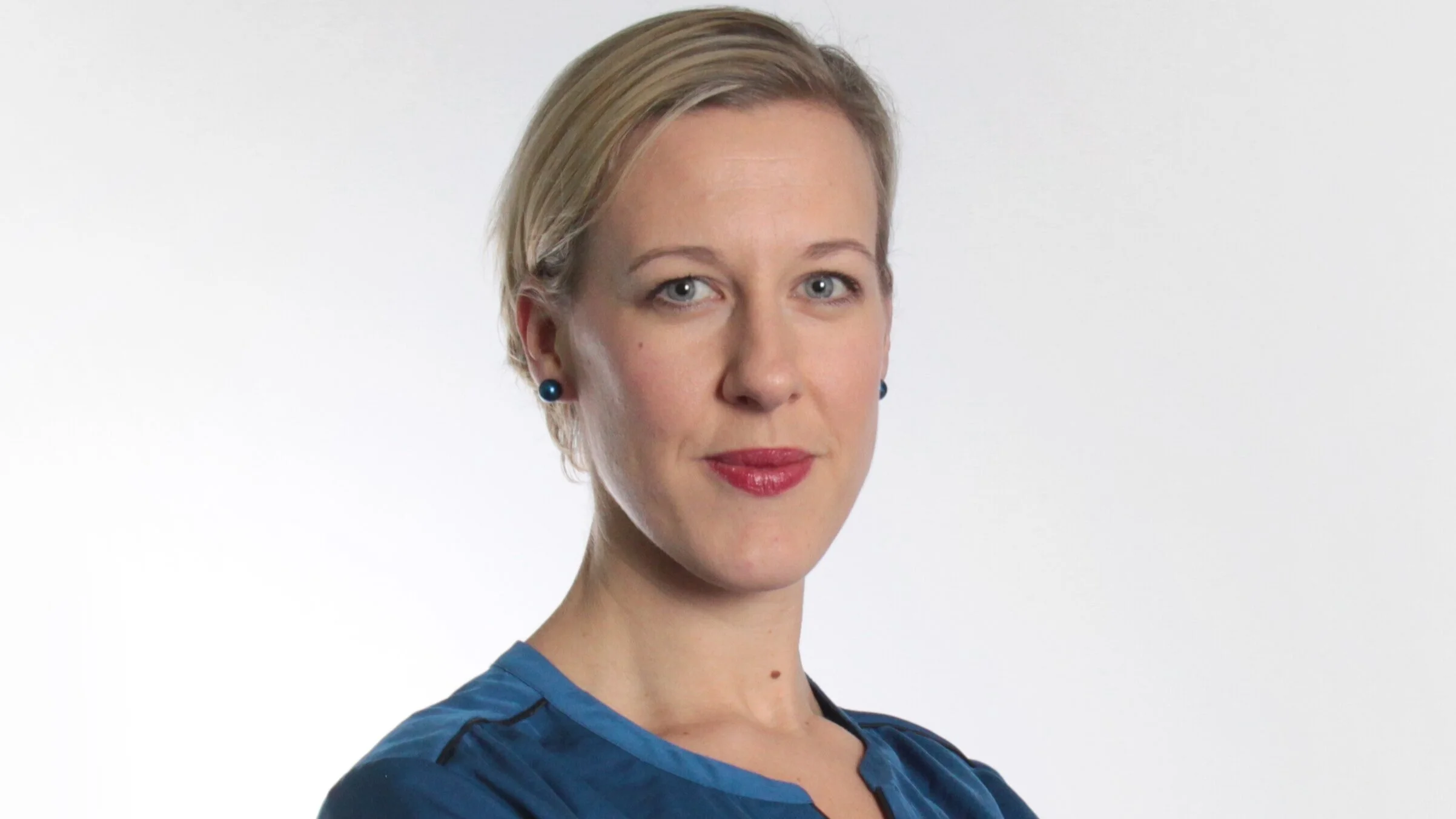 Porträt einer Frau mit blonden kurzen Haaren, blauen Augen und rotem Lippenstift, trägt eine blaue Bluse, vor weißem Hintergrund.