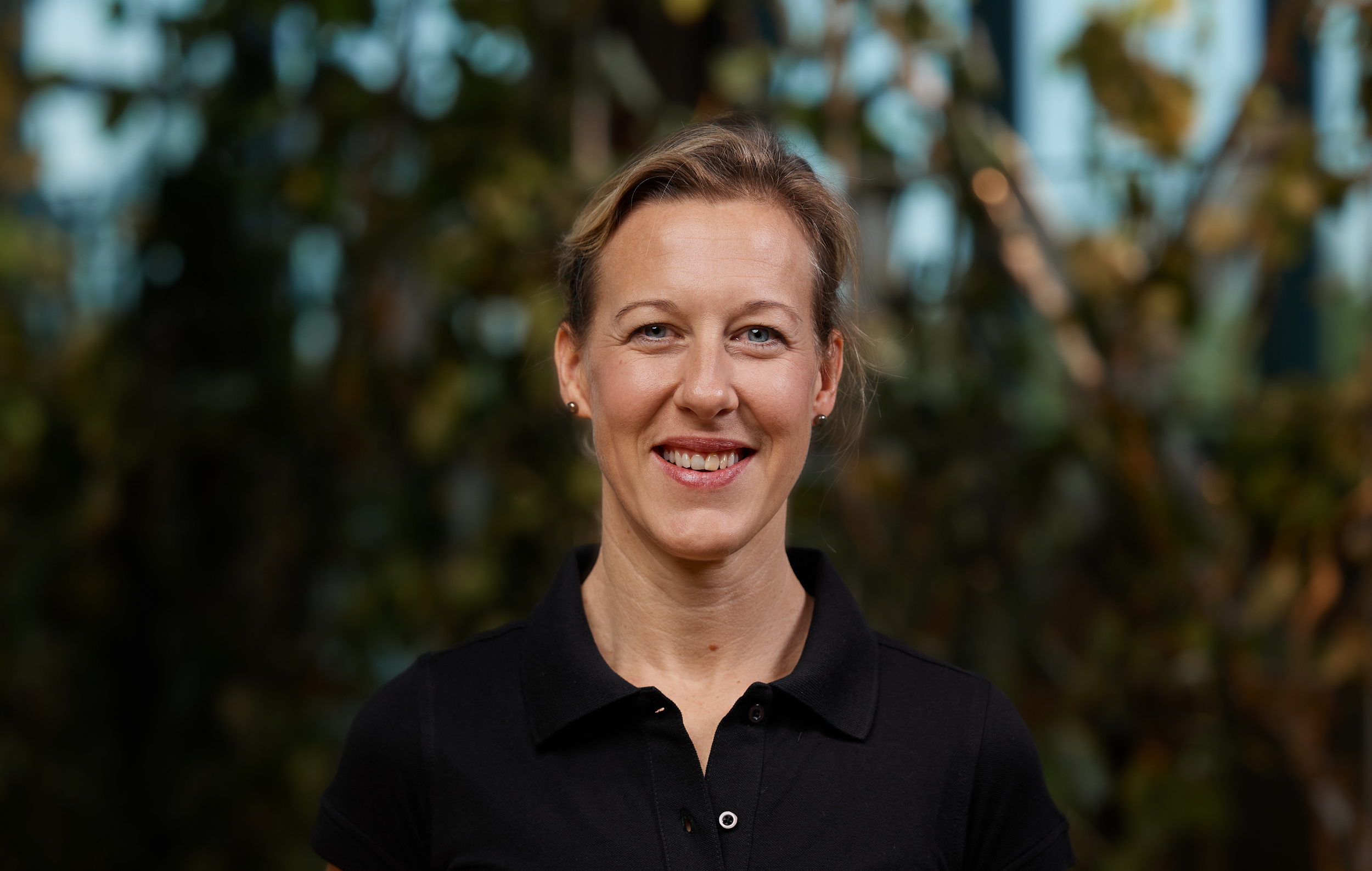 Porträt einer lächelnden Frau mit kurzen, hellen Haaren, trägt ein schwarzes Polohemd, im Hintergrund unscharfe grüne Blätter und Äste.