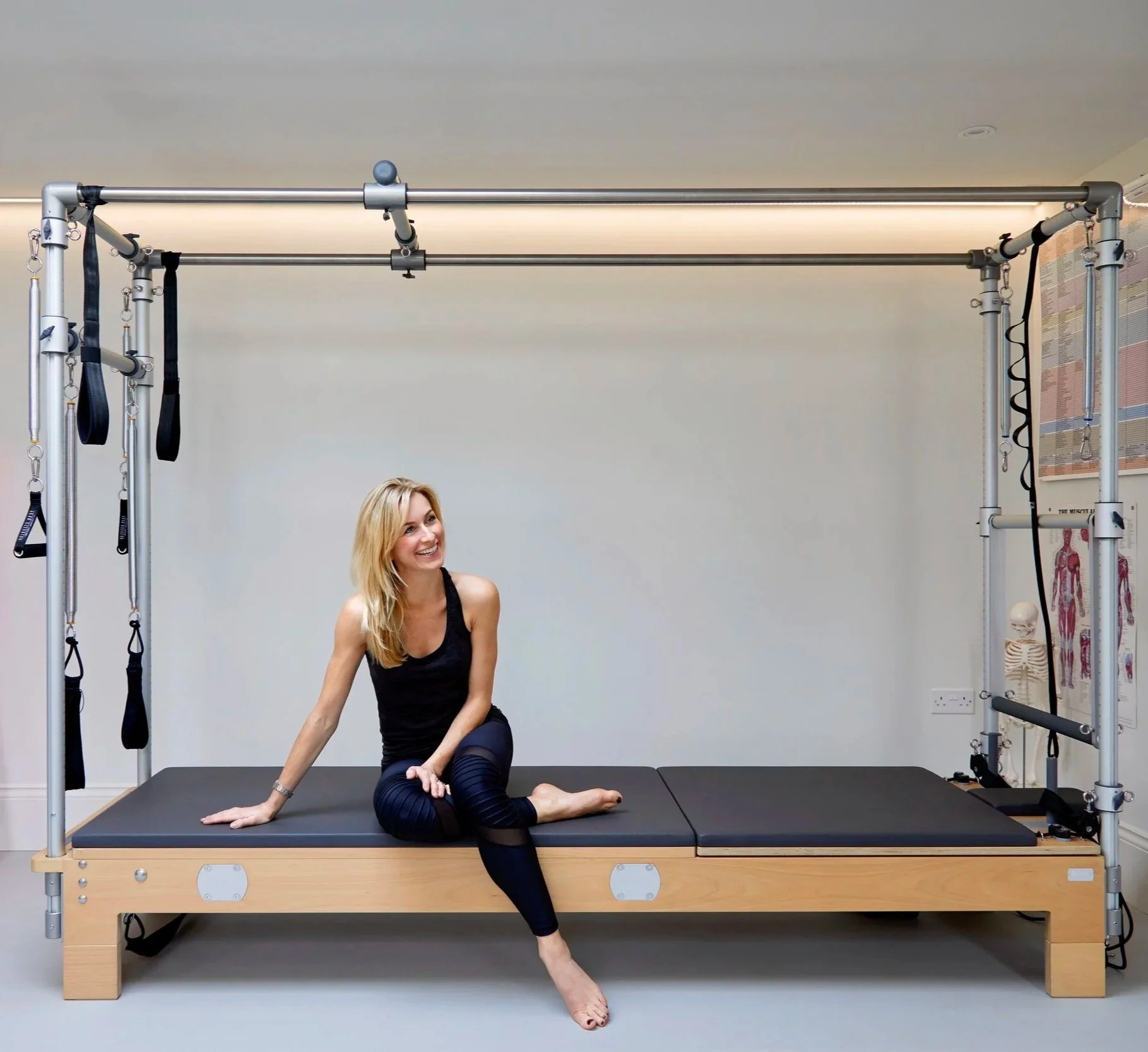 Emily Odoire sitting on a Pilates cadillac machine in a fitness studio, smiling and looking to the side.