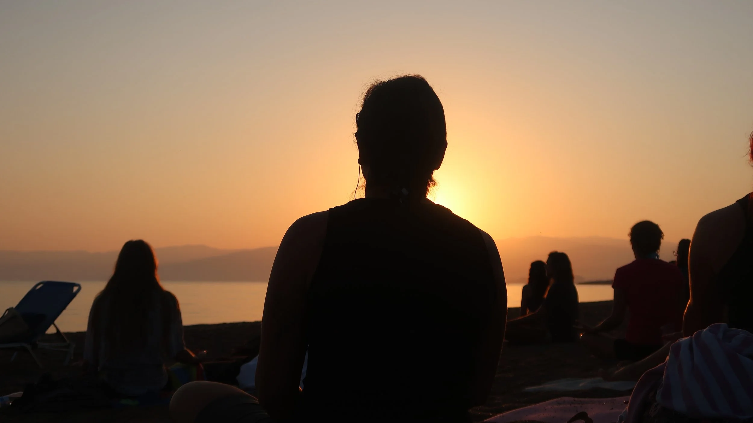 Silhouettes of people sitting on a beach during sunset, facing the water and mountains in the distance.