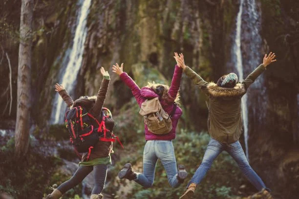 Three people jumping with arms raised in a forest near a waterfall.