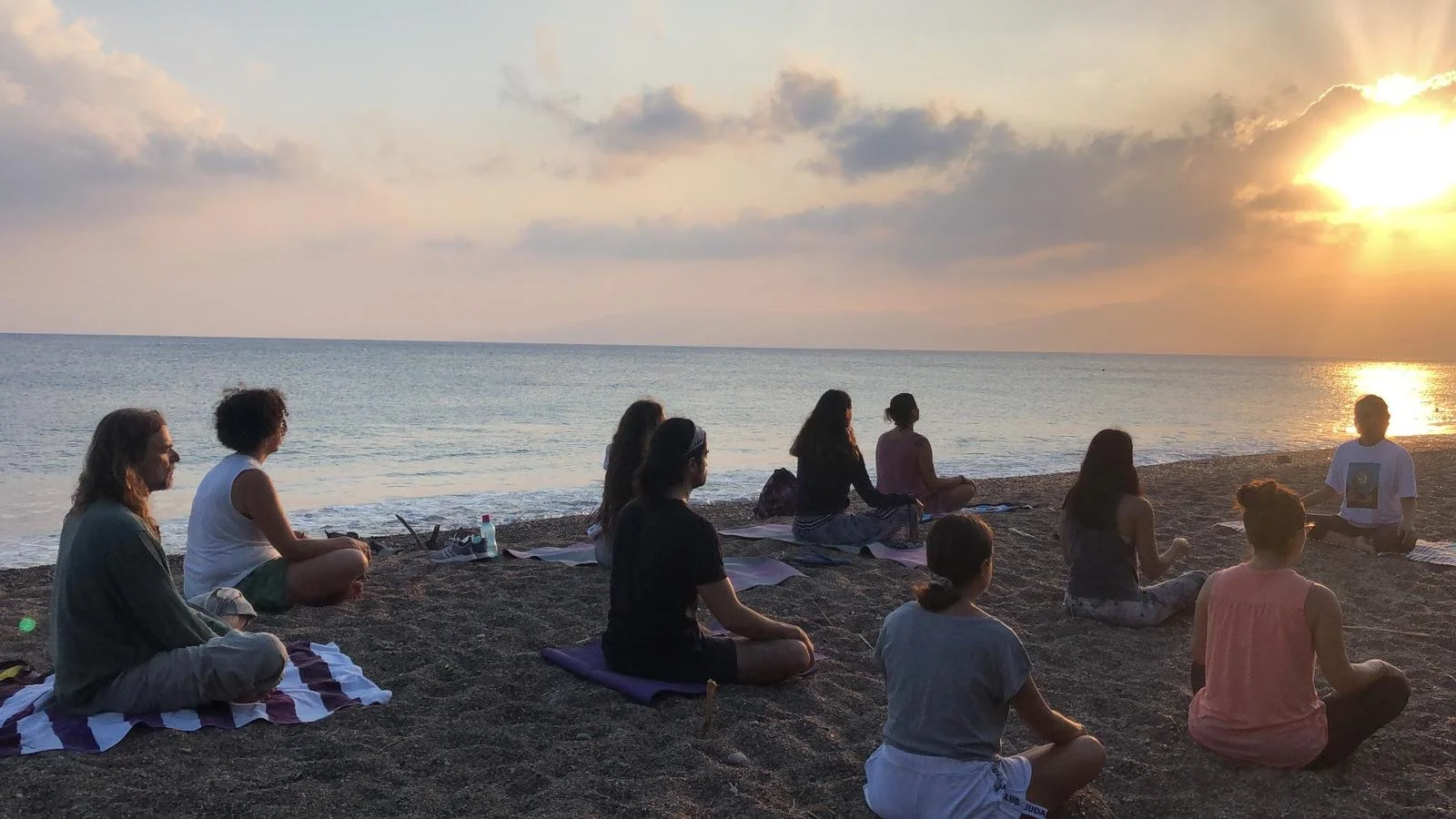 A group of people sitting on a sandy beach during sunset, practicing meditation or yoga near the water.