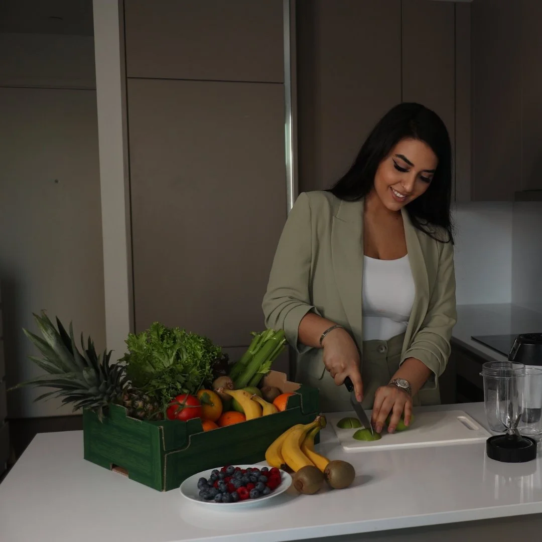 A woman cutting a lime in a modern kitchen with a variety of fresh fruits and vegetables on the counter, including pineapple, bananas, kiwis, blueberries, raspberries, grapes, and a box of assorted produce.
