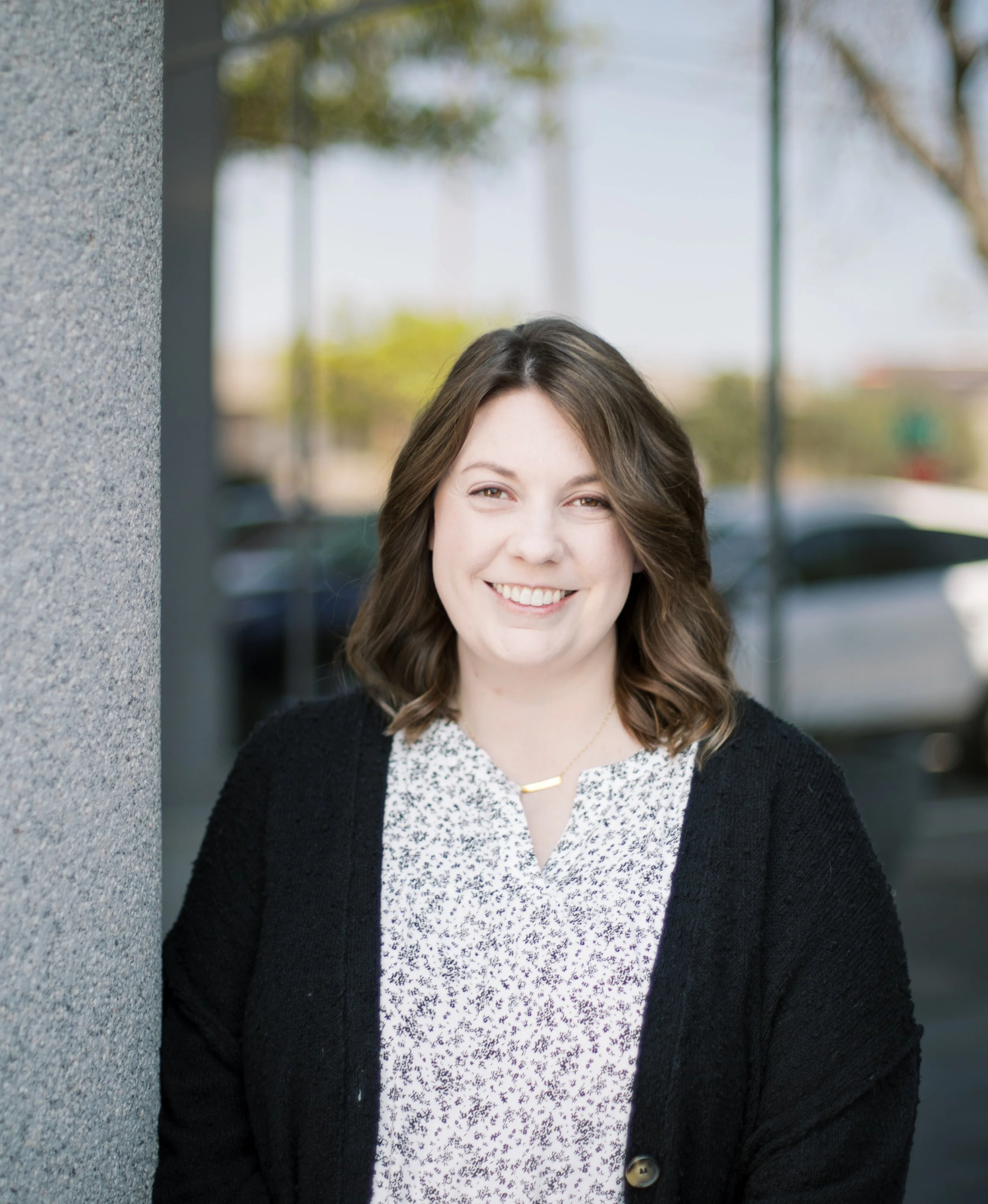 Smiling woman with shoulder-length brown hair, wearing a black cardigan over a white patterned top, standing outdoors in front of a blurred background with trees.