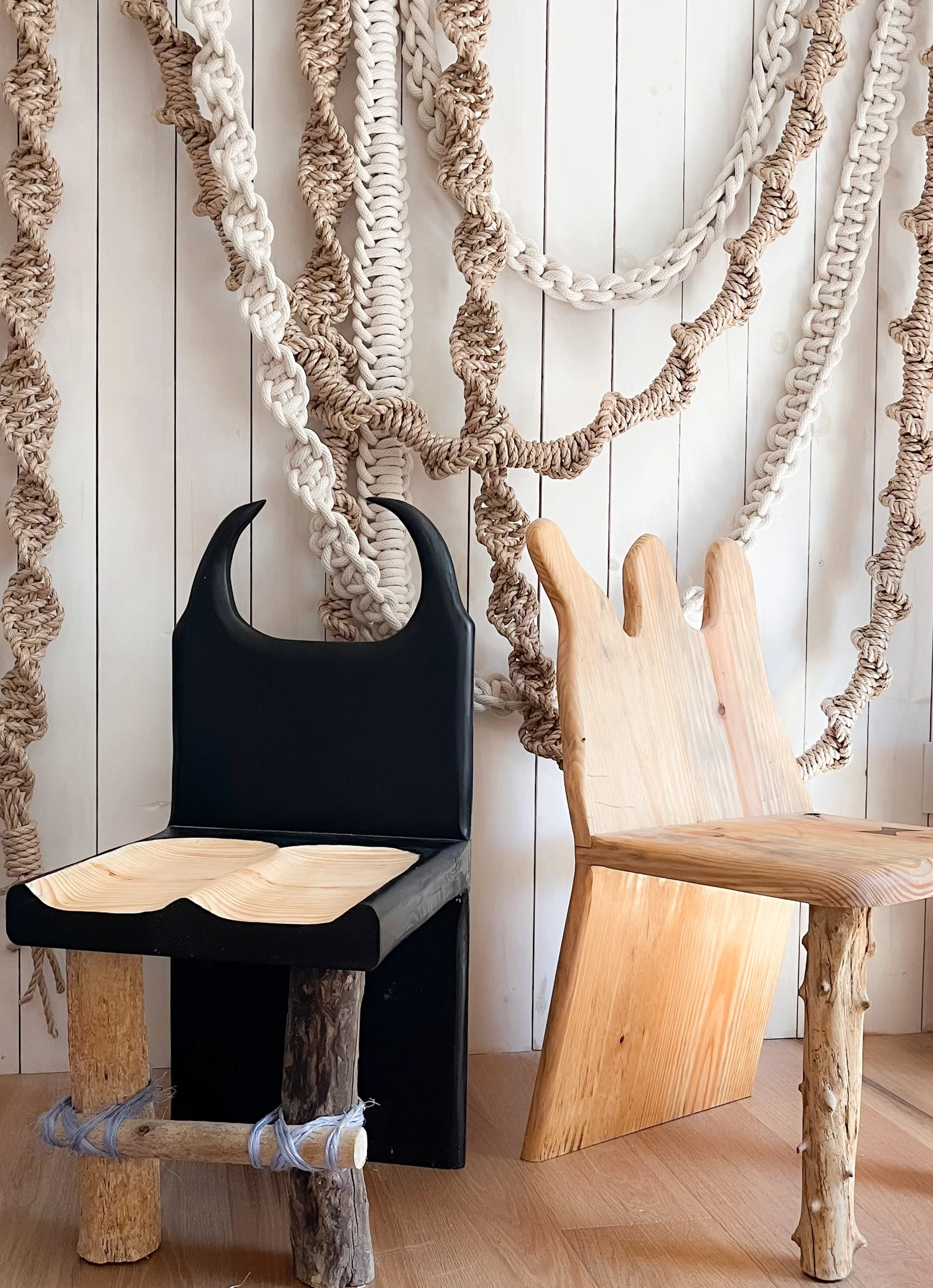 Two chairs, one black and one natural wood, with woven rope and wood accents, set against a white wooden wall decorated with large knotted ropes.