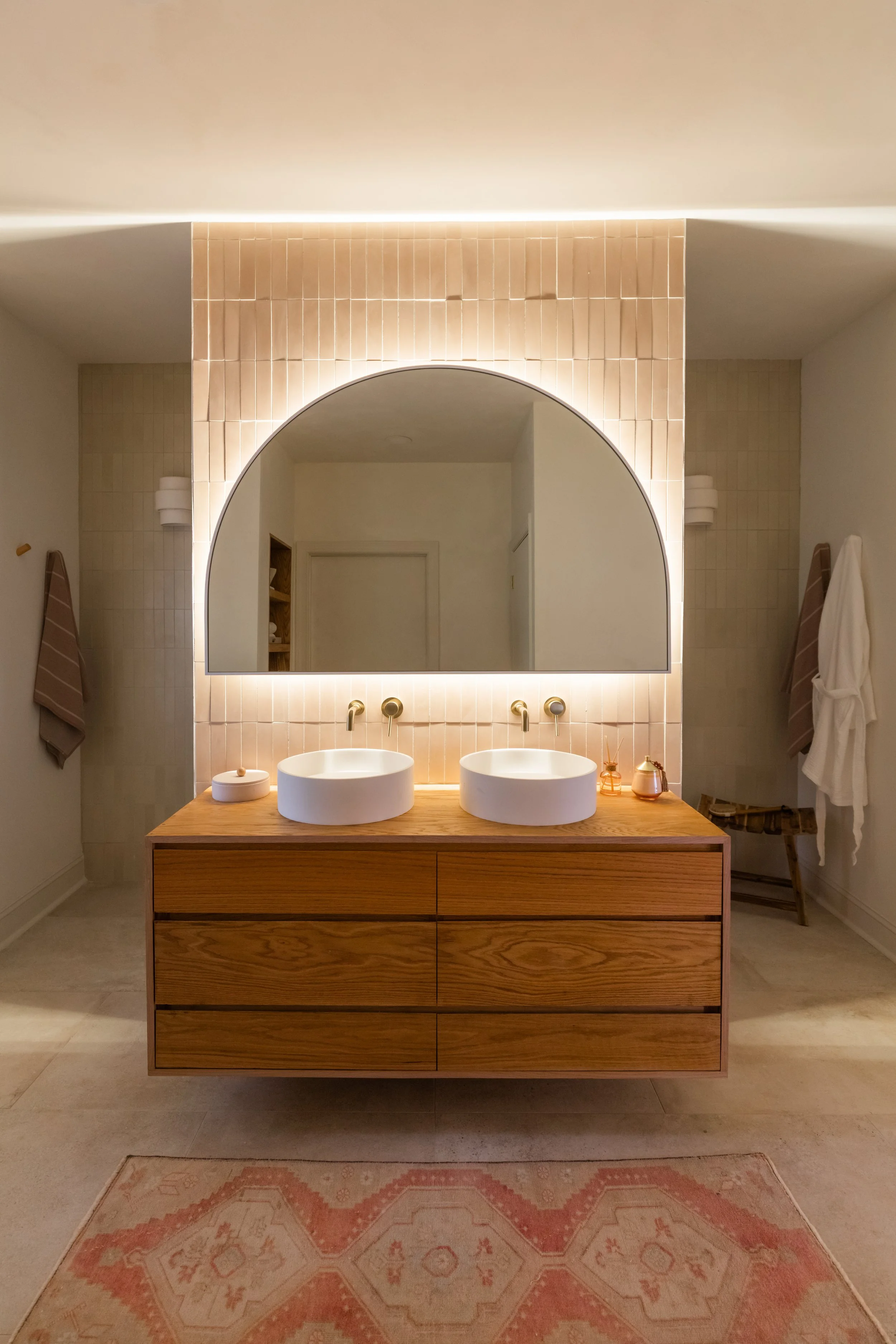 Modern bathroom with a wooden double vanity, two round vessel sinks, large mirror with backlighting, beige tiled walls, wall-mounted faucets, and soft lighting. Towels and decorative items are on the side.
