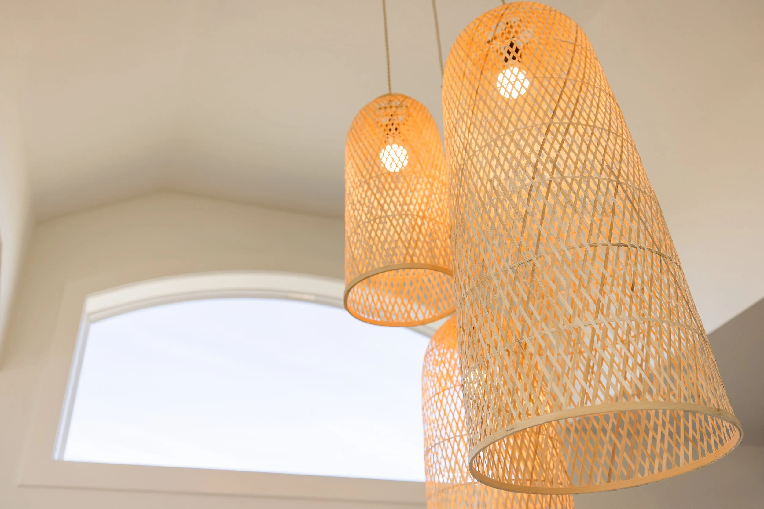 Close-up of three woven rattan pendant lights hanging from the ceiling near a window with a white arch.