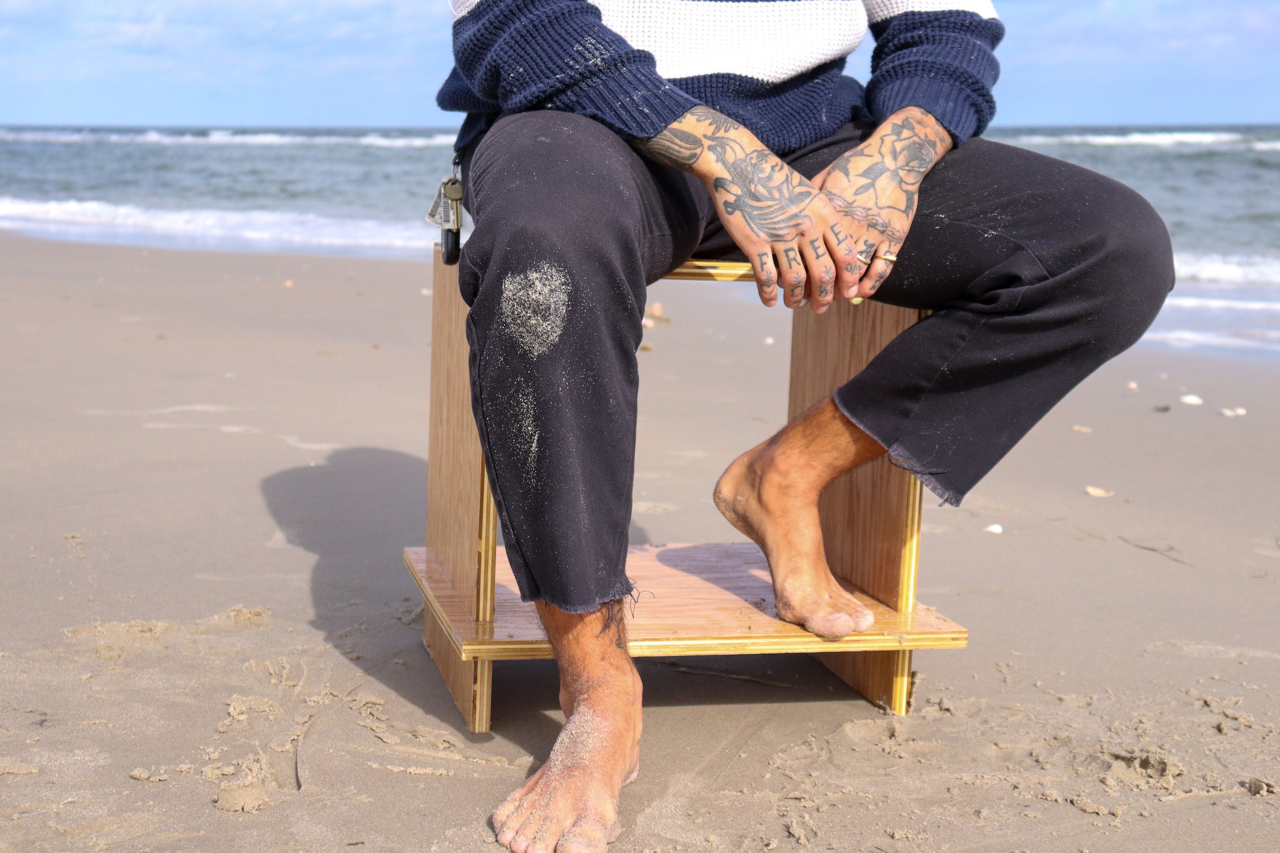 Person sitting on a small wooden rectangular structure on a sandy beach with ocean waves in the background. The person is barefoot, with sand on their legs and feet, wearing black shorts and a long-sleeved shirt. They have tattoos on their hands and 