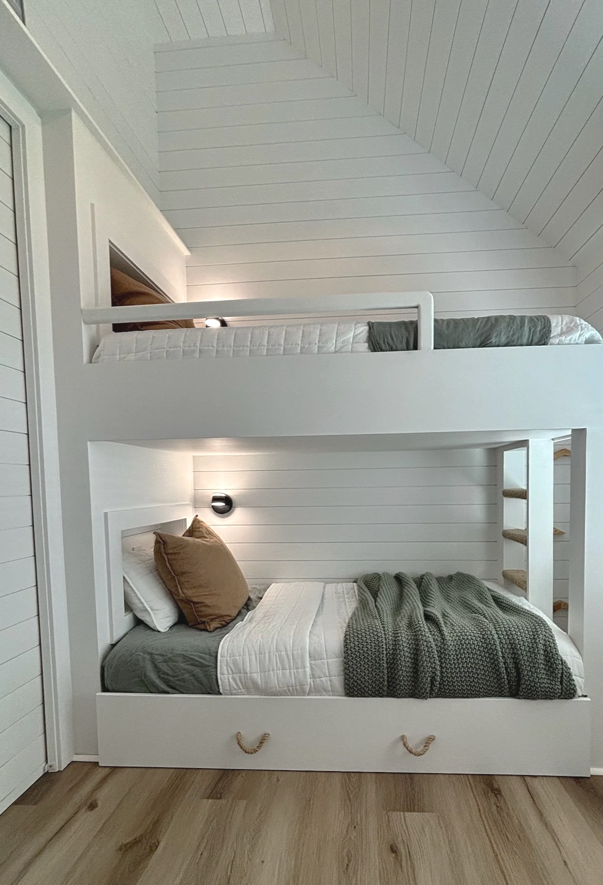 A bedroom with a white bunk bed, the lower bed is made with a gray blanket and brown pillows, and the upper bed has a white mattress and gray pillows. The room features white wood-paneled walls and ceiling, and a corner light fixture near the ceiling