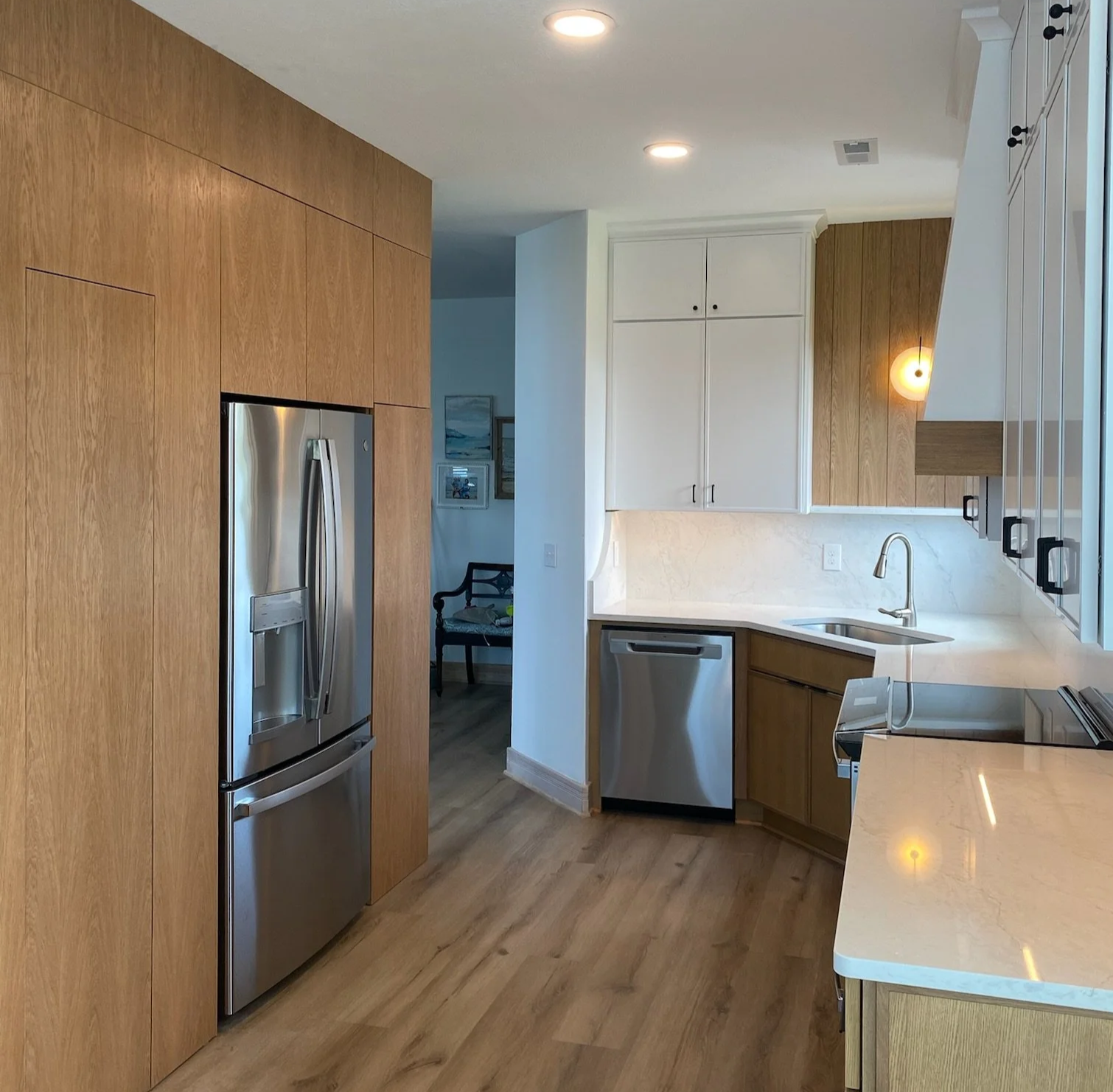 Modern kitchen with wooden and white cabinets, stainless steel refrigerator and dishwasher, beige marble countertops, and hardwood floors, with recessed lighting.