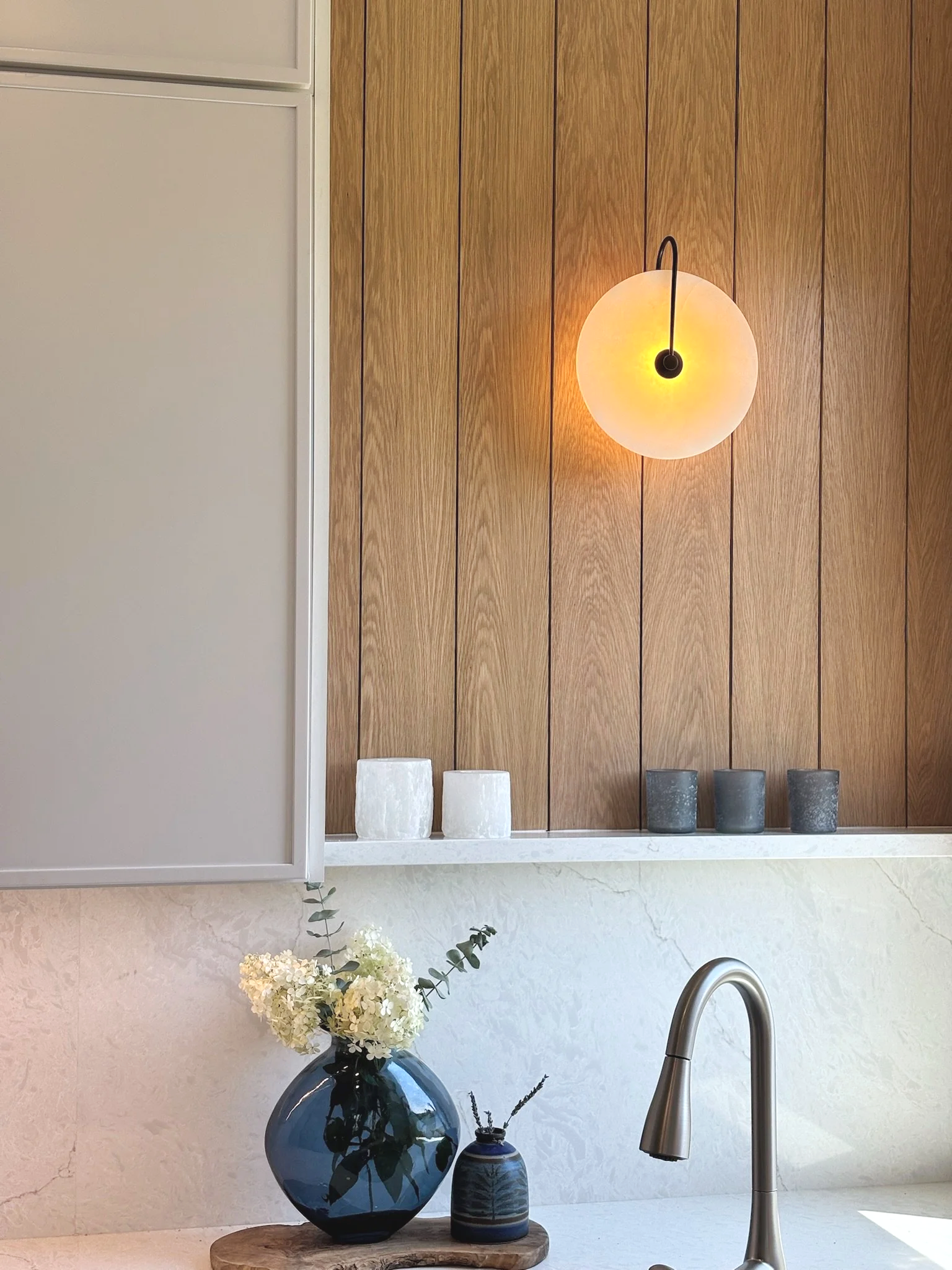 A kitchen interior with a wooden wall, white cabinet, modern metallic faucet, a large dark blue vase with white flowers on a wooden surface, and three smaller decorative vases nearby.