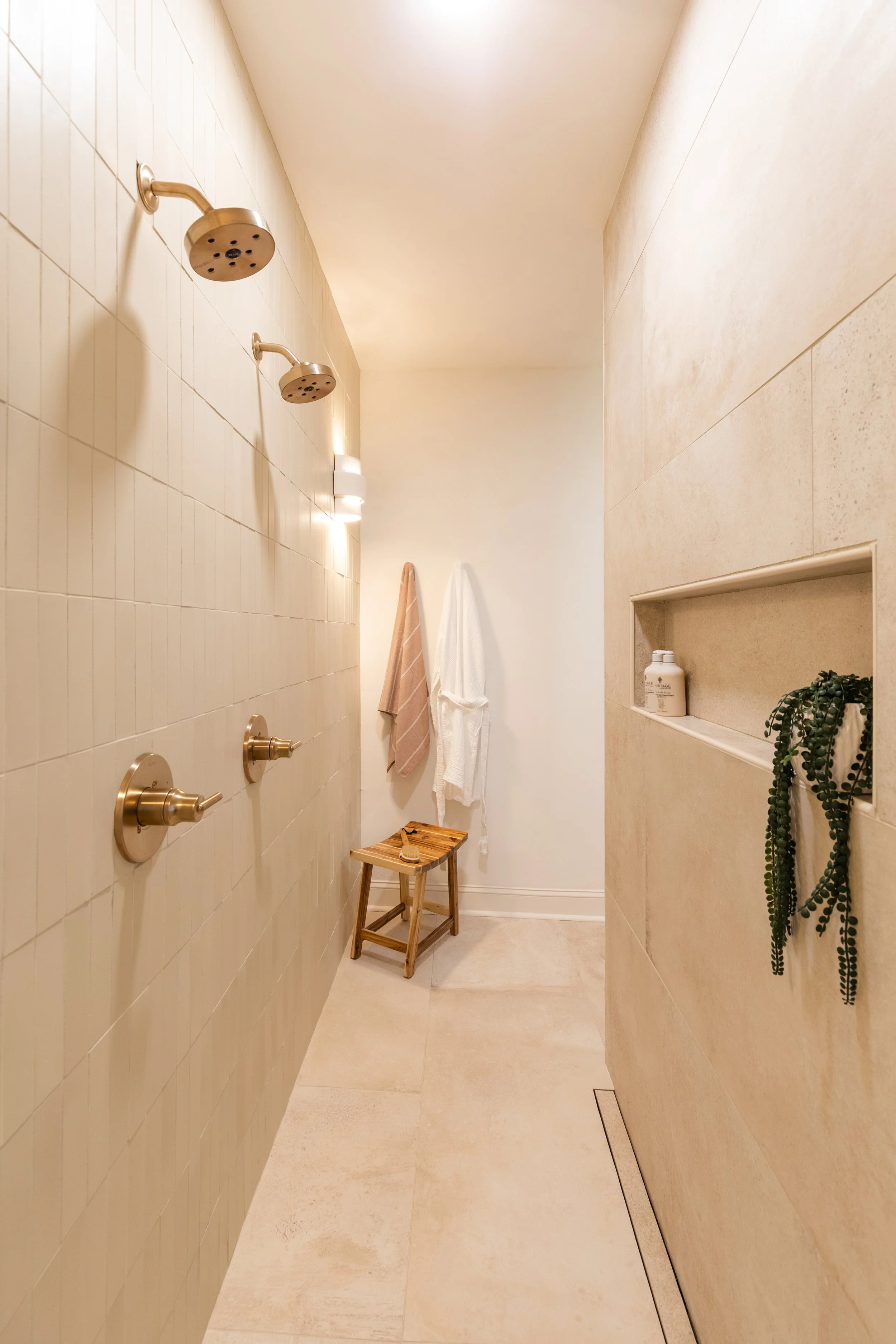 A minimalist bathroom with cream-colored tiled walls and floor. There are two rainfall showerheads on the left wall, with three bronze shower controls below them. Towels are hanging on the back wall, and a small wooden stool is in the corner. Built-i