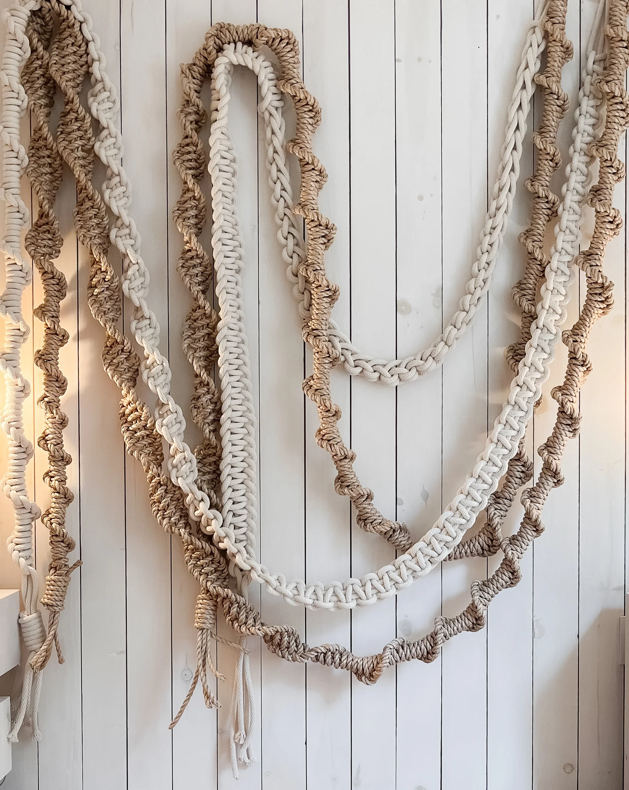 Multiple thick, twisted ropes in natural and cream colors hanging on a white wooden wall.