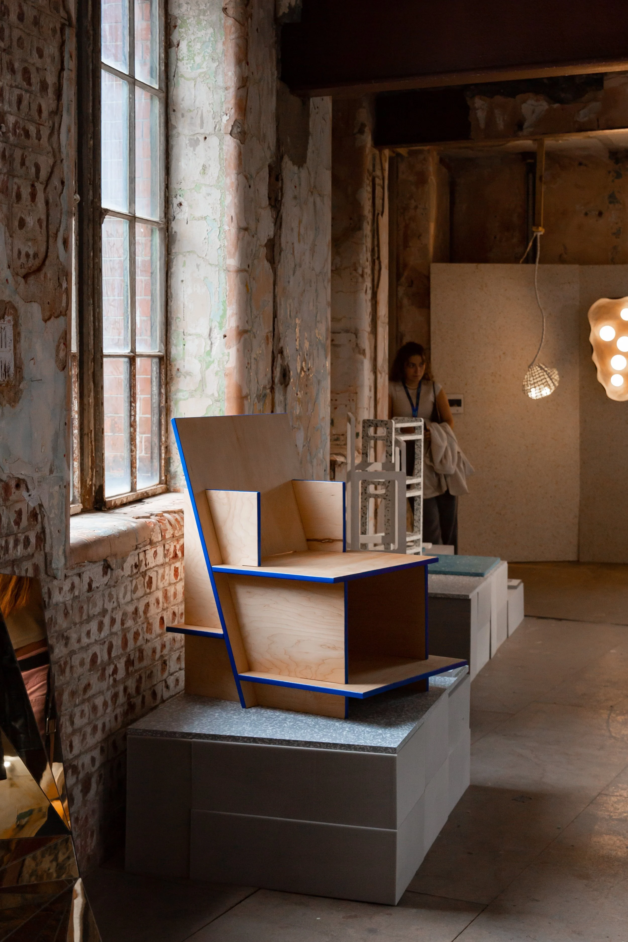 A display of modern, unfinished wooden furniture in a brick-walled interior with large windows at an art gallery show. The furniture includes a stepped shelving unit with a blue border and a grey cabinet below. 