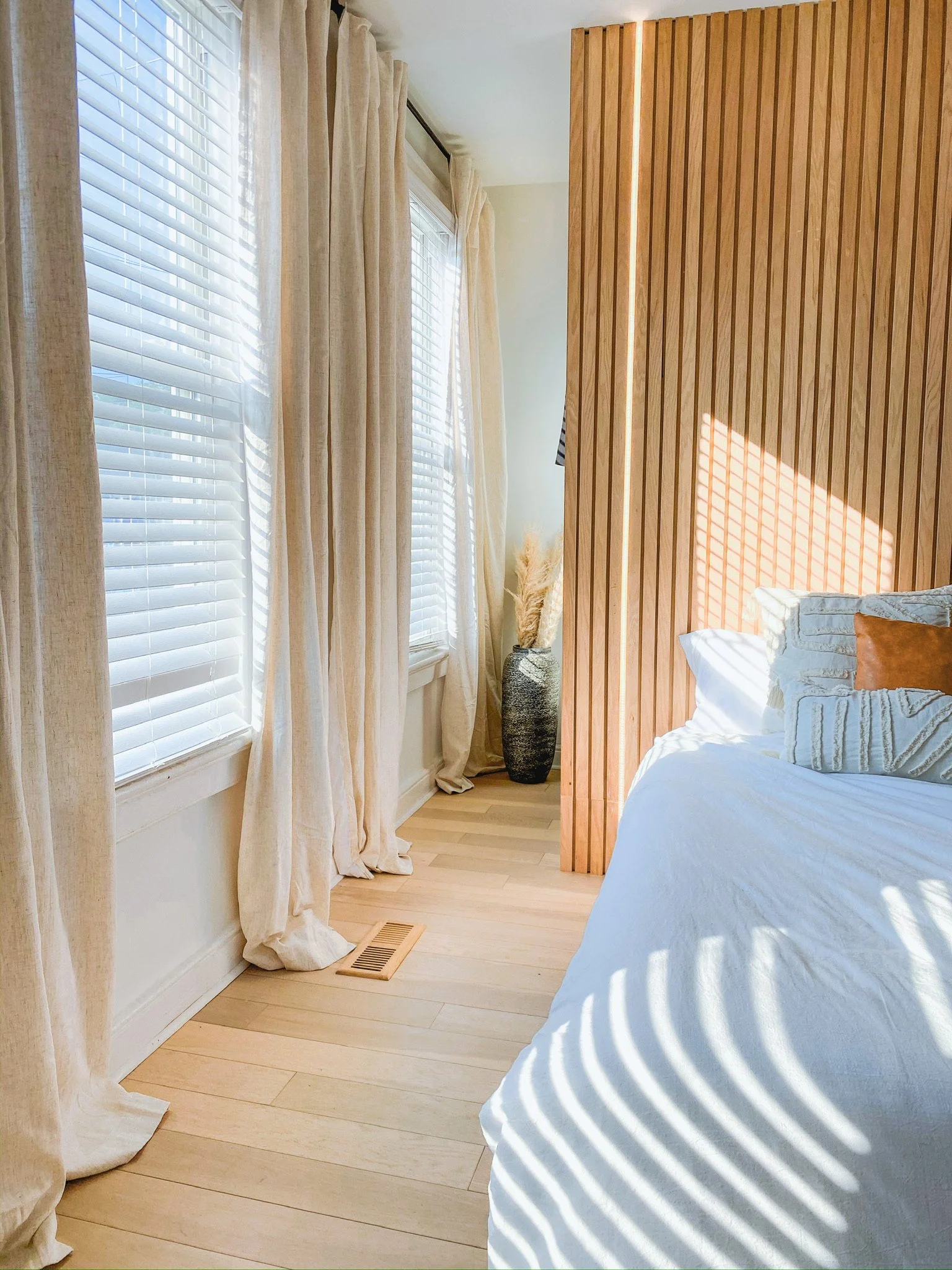 A corner of a bedroom with closed white blinds on windows, beige curtains, a wooden floor, a tall ceramic vase with pampas grass, and part of a bed with white bedding and a brown pillow, illuminated by sunlight creating shadows.
