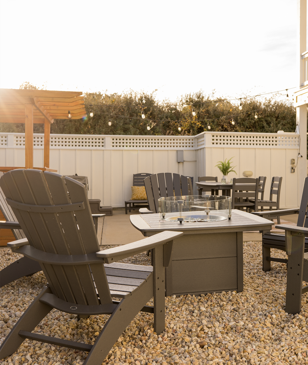 Outdoor patio with Adirondack chairs around a fire pit, string lights overhead, and a raised dining table with chairs and a potted plant.