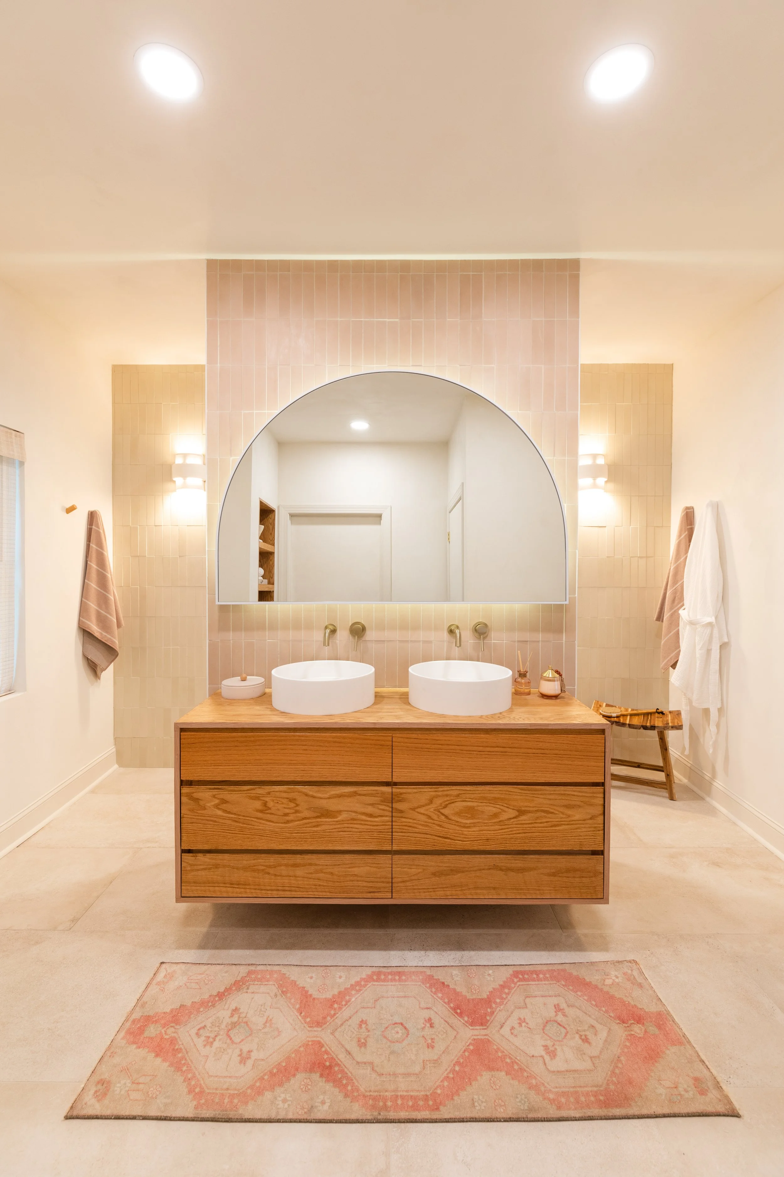 Bathroom with two round vessel sinks on a wooden vanity, large mirror above, pink tiled wall, beige walls, towels hanging on the wall, pink patterned rug on the tile floor.
