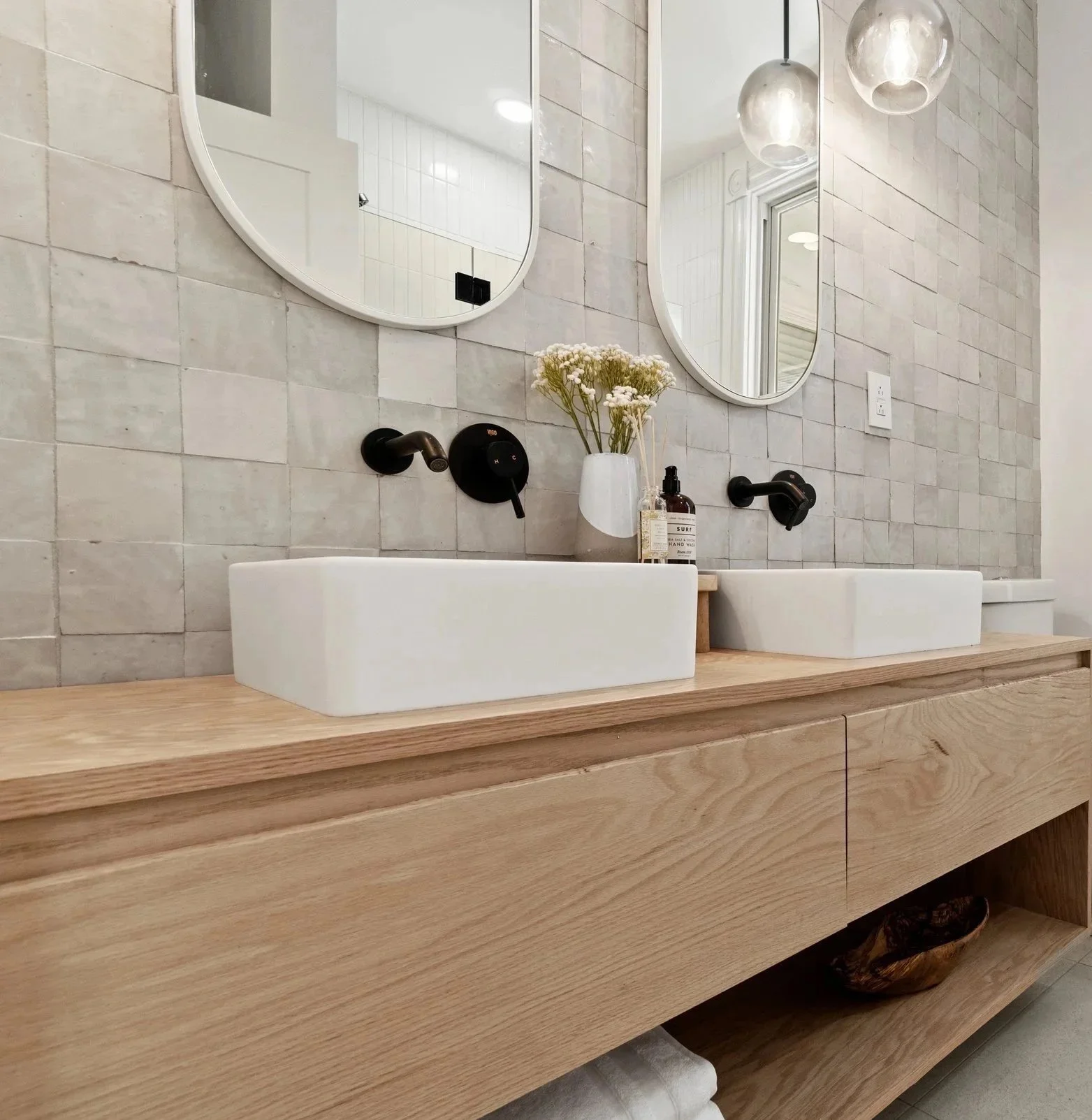 A modern bathroom sink vanity with two white vessel sinks, black wall-mounted faucets, two oval mirrors, a vase with white flowers, and a mirror-backed wall with light fixtures.