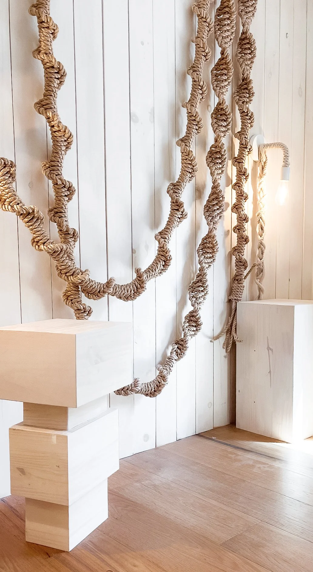 Decorative beige rope hanging in loops on a white wooden plank wall, with sculptural white wooden nightstands and a wall sconce light fixture to the right.