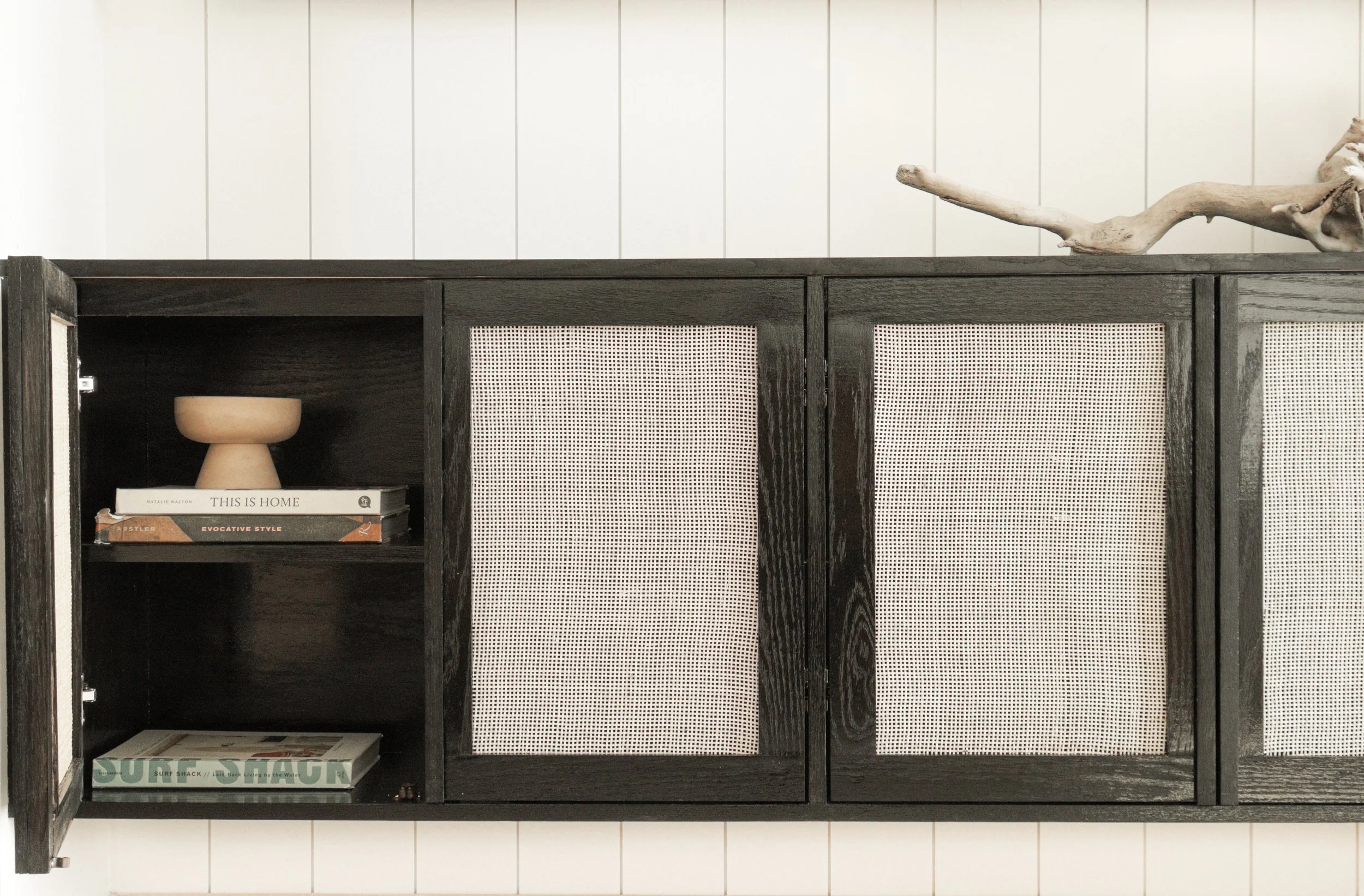 A black wooden cabinet with two frosted glass panels, a small shelf containing books and a decorative bowl, and an open section with a book titled "This Is Home" on the bottom shelf. There is a large piece of driftwood on top of the cabinet, set agai
