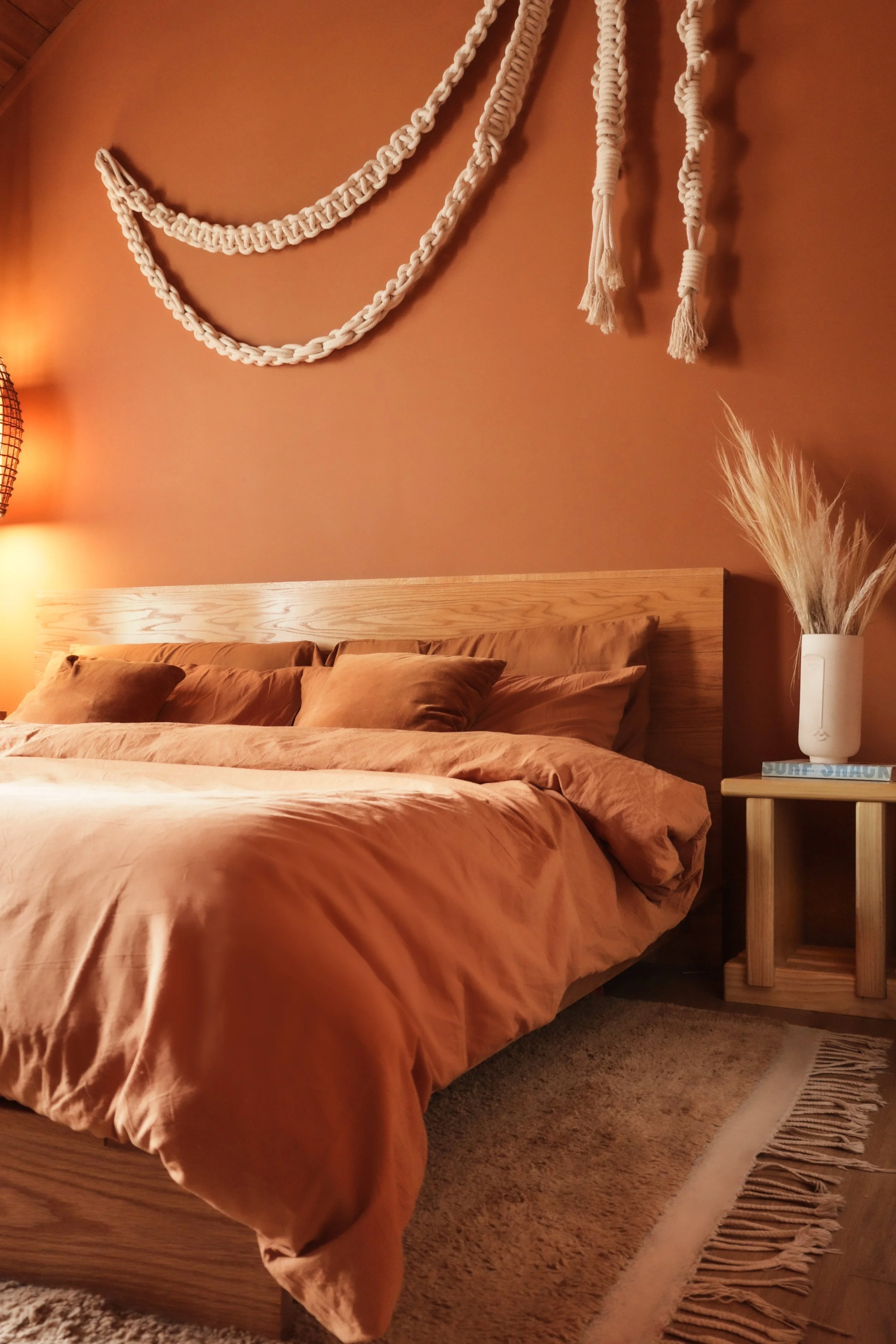 A cozy bedroom with warm lighting, featuring a bed with  rust and terracotta-colored duvet and matching pillows, a wooden headboard, a beige moroccan rug with fringes, and decorative cream-colored knotted wall hangings.