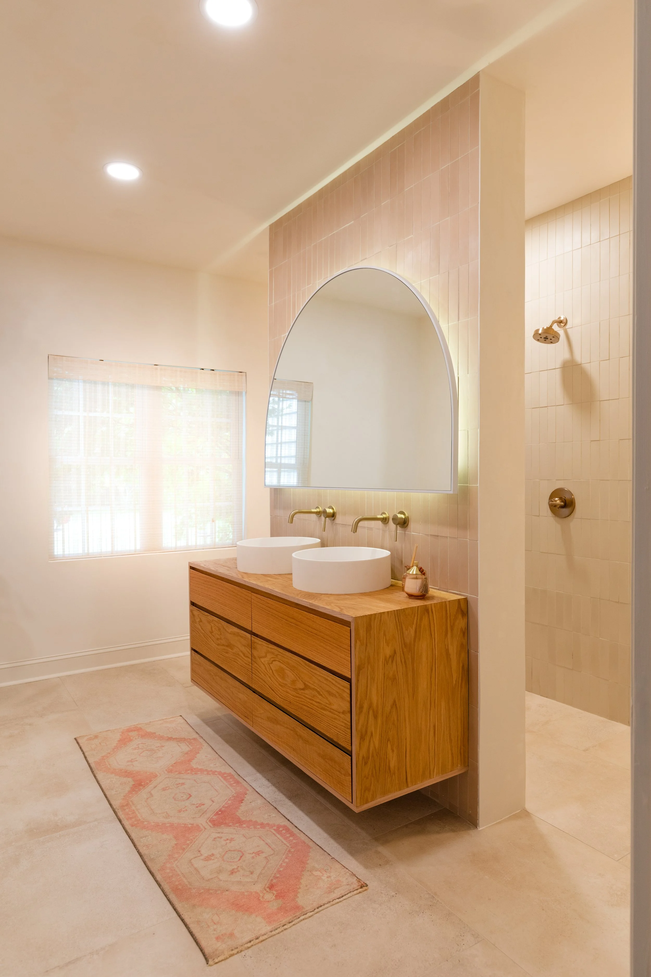 A bathroom with a wooden vanity with two white vessel sinks, brass faucets, a large arched mirror, a pink tile wall behind the mirror, a window with blinds, a shower with beige tiles and a rain showerhead, and a pastel pink patterned rug on beige til