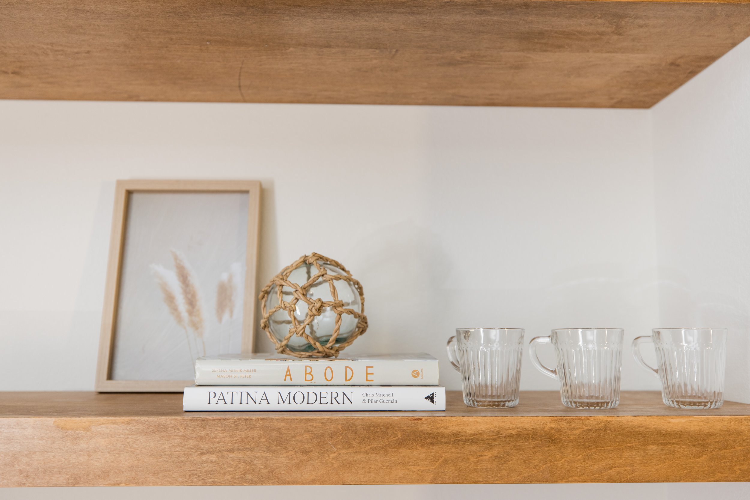 A wooden shelf displaying three books, a decorative wicker ball, and three glass cups; a framed art piece leans against the wall behind the books.