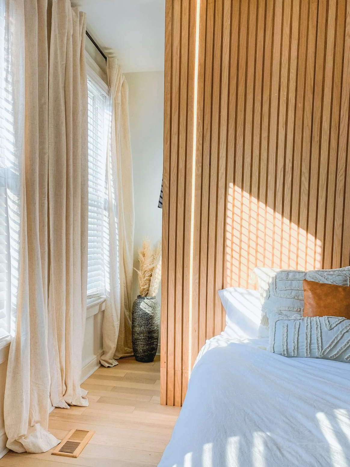 A bedroom with cream curtains, a tall vase with pampas grass, a wooden slat wall, part of a bed with white linens and a brown pillow, and sunlight creating shadows through window blinds.