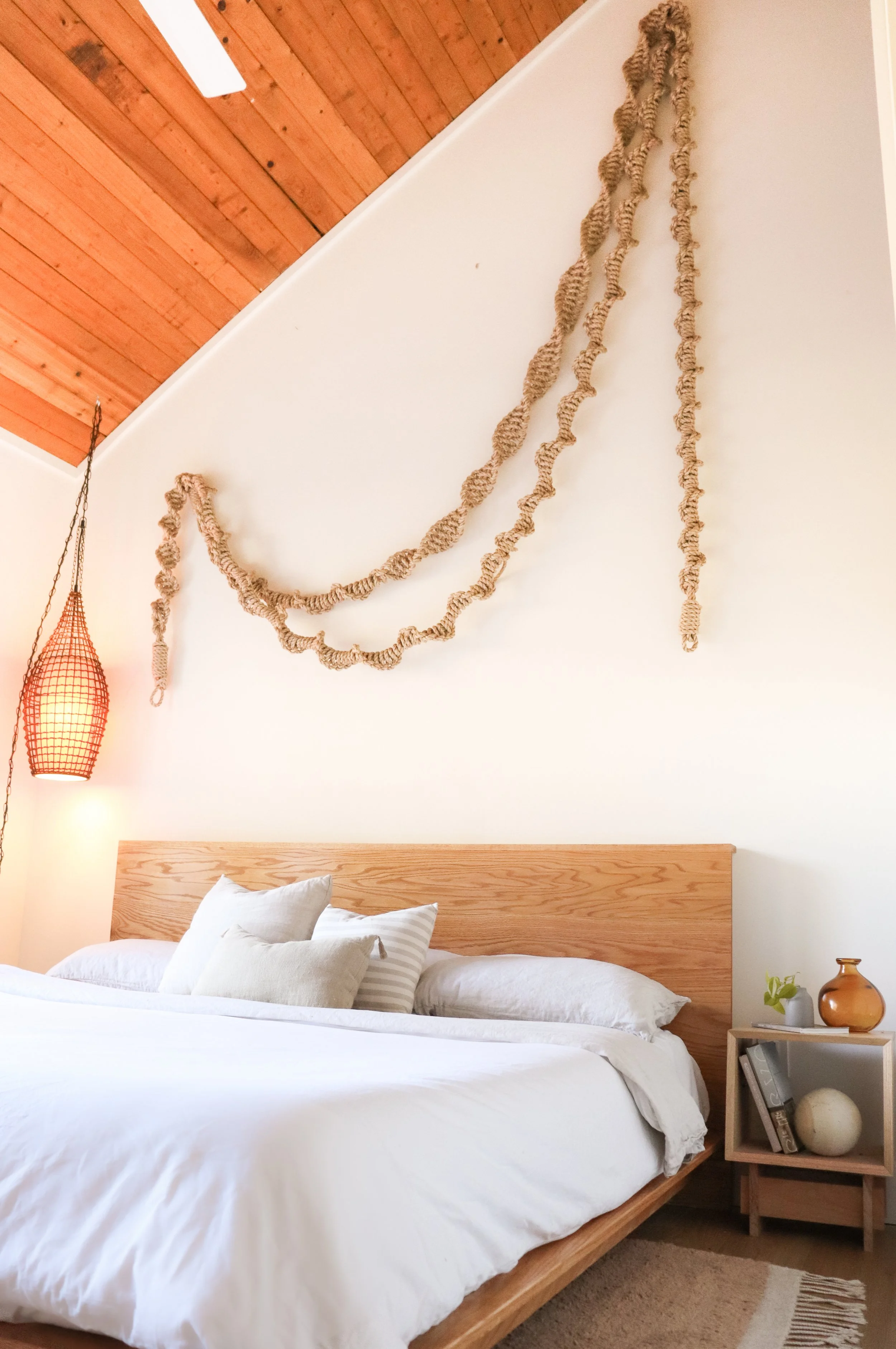 A cozy bedroom with a wooden bed frame and white bedding including pillows. A nightstand with decorative items is beside the bed, and a woven pendant light hangs in the corner. The wall behind the bed has a creamy color with a wall hanging made of tw