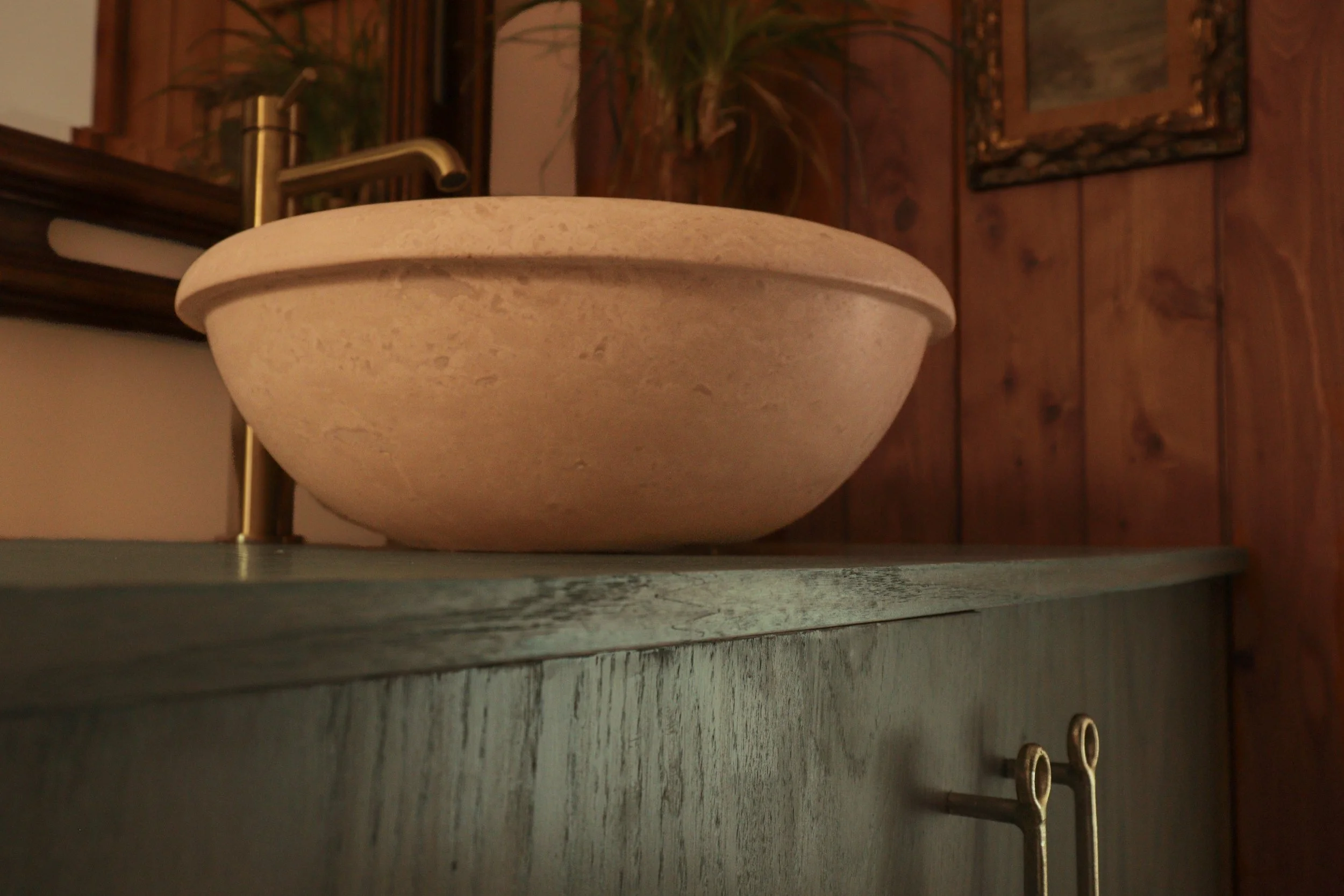 Close-up of a beige round ceramic basin on a countertop in a rustic wooden interior, with a brass faucet, potted plant, framed artwork, and two rings on a string in the background.