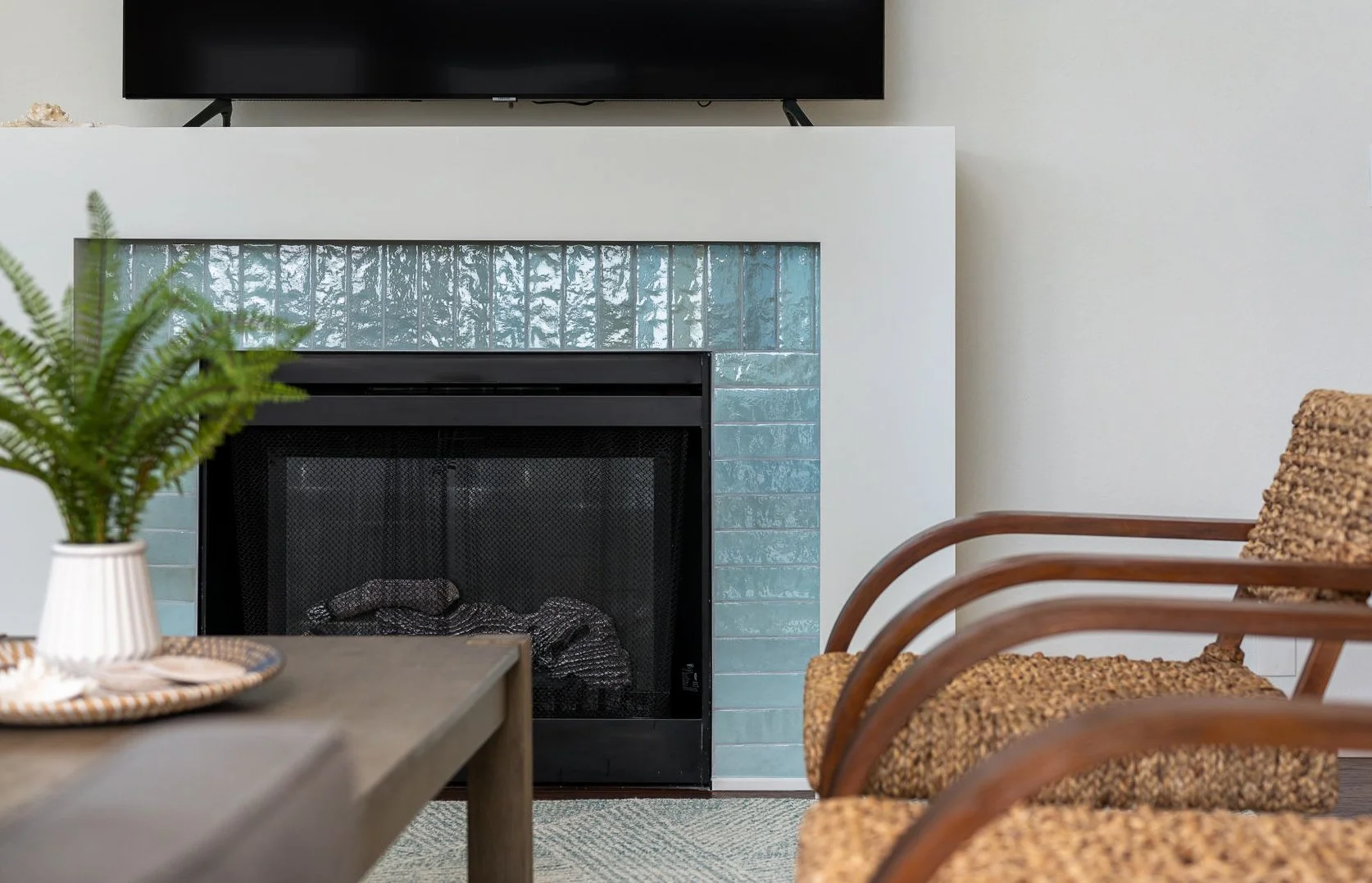 Living room with a fireplace surrounded by blue tiles, a mounted flat-screen TV above, a wooden chair with a patterned cushion, a coffee table with a potted plant, and part of a woven basket in a neutral-colored room.