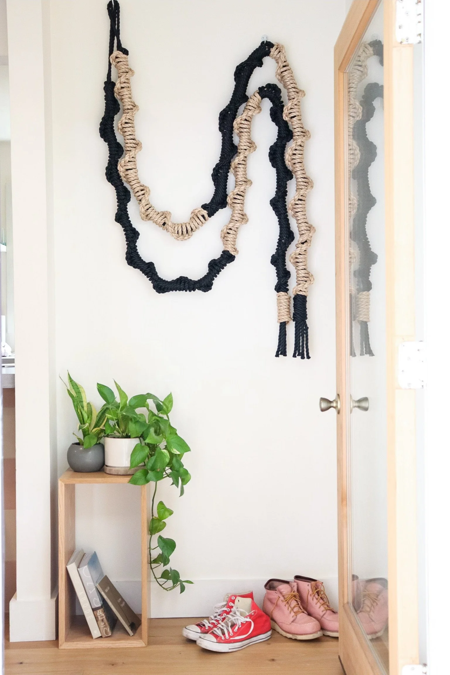 Entryway with floating shelves holding books and a potted plant, a mirror reflecting the hanging wall art, and shoes on the floor including red sneakers and pink boots.