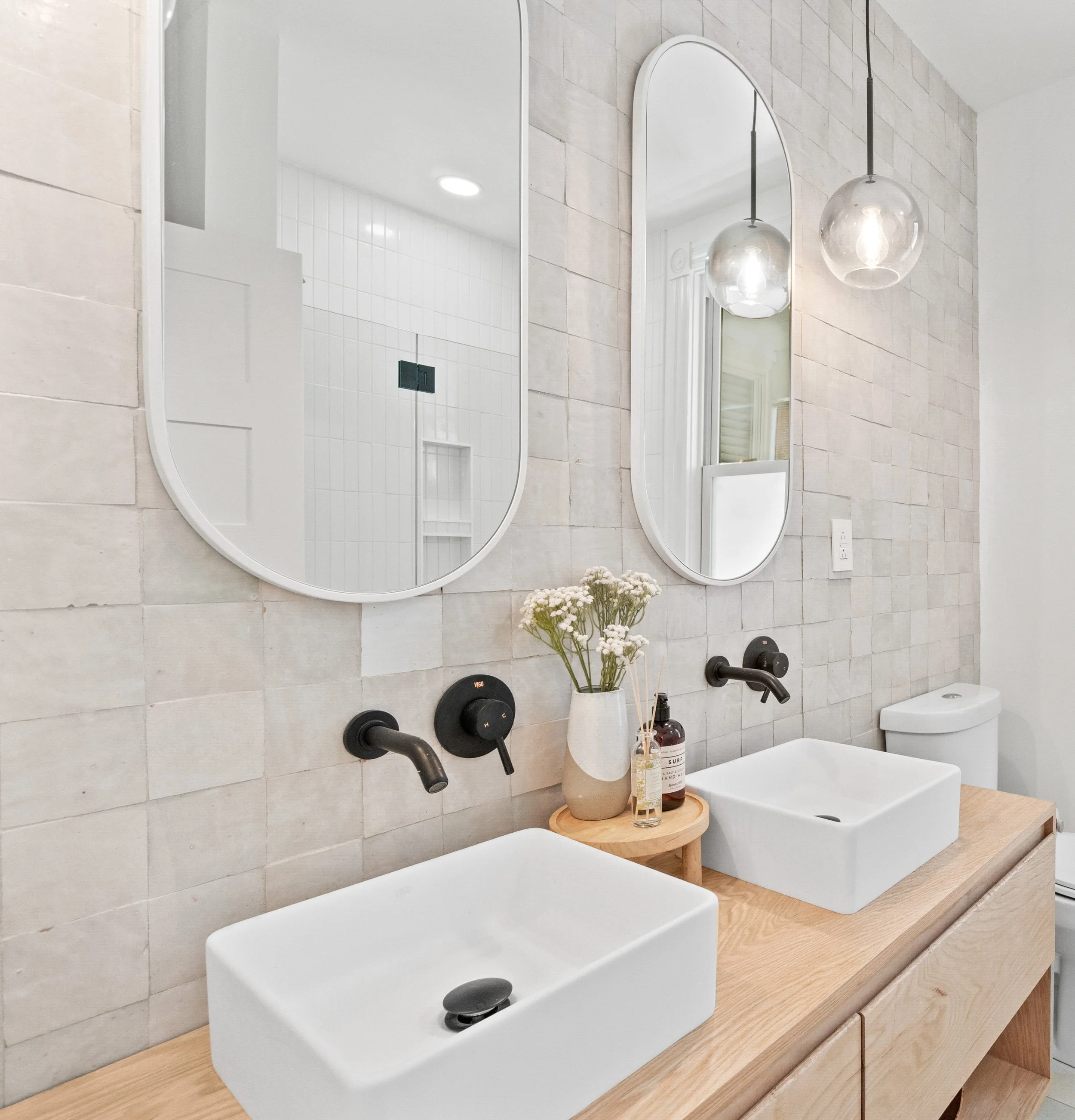 Modern bathroom with two vessel sinks on a wooden vanity, black wall-mounted faucets, large oval wall mirrors, decorative flowers in a white vase, and hanging glass globe pendant lights.