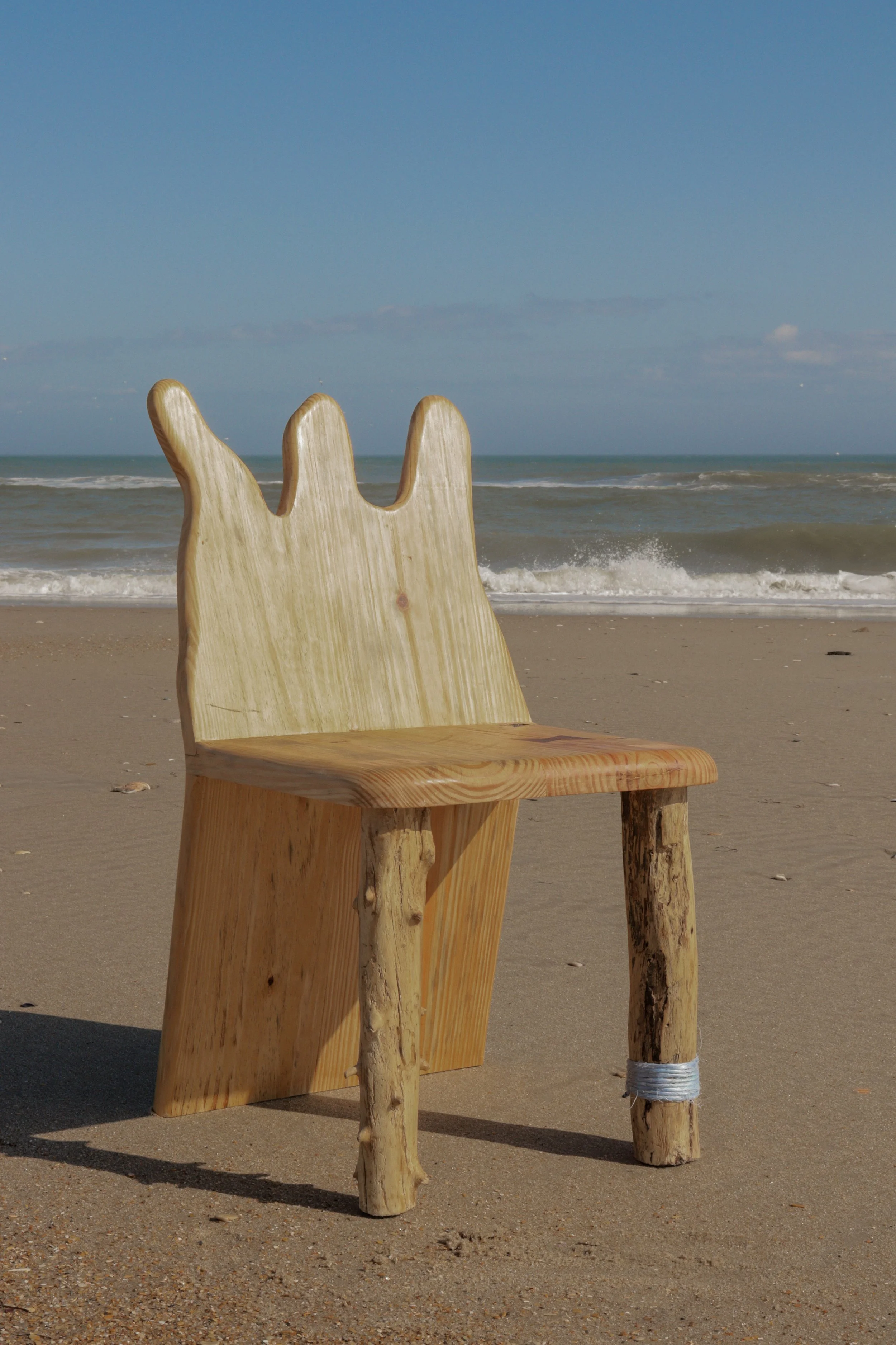 Wooden chair with backrest resembling a hand, situated on a sandy beach near the ocean with a blue sky overhead.