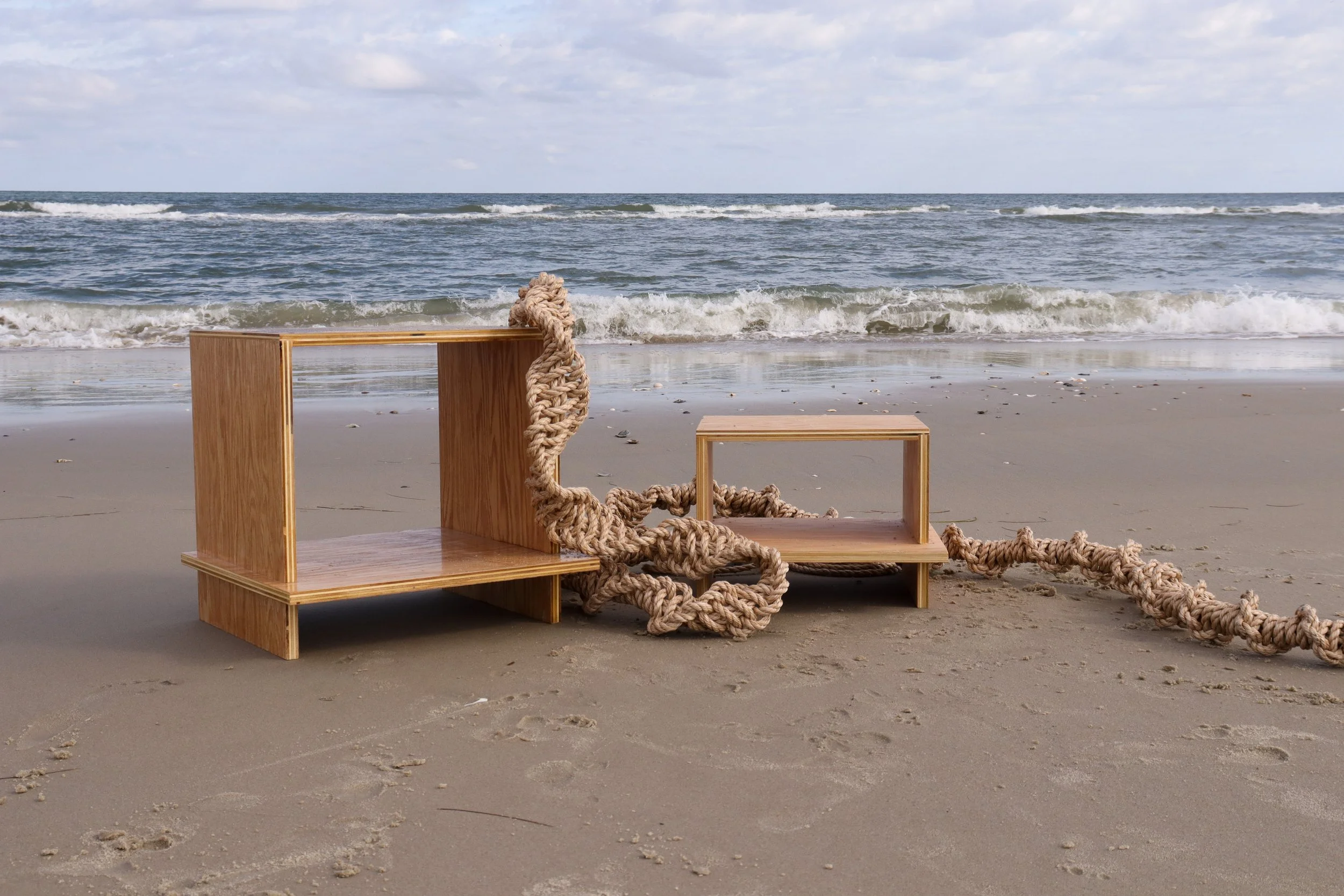 Wooden furniture pieces with thick woven ropes on a sandy beach facing the ocean under a cloudy sky.