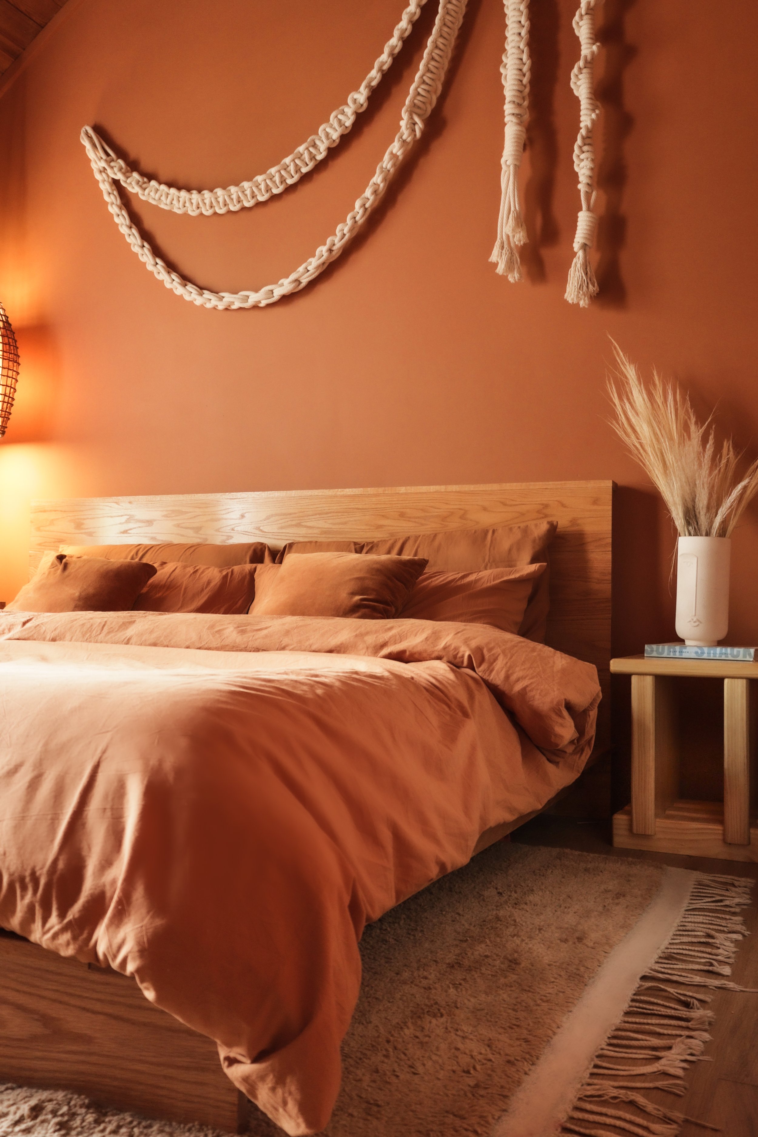 A bedroom with a wooden bed, beige bedding, and pillows. Decor includes three white braided wall hangings on an ochre-colored wall and a white vase with pampas grass on a small wooden nightstand. There is a beige rug with fringes on a carpeted floor.