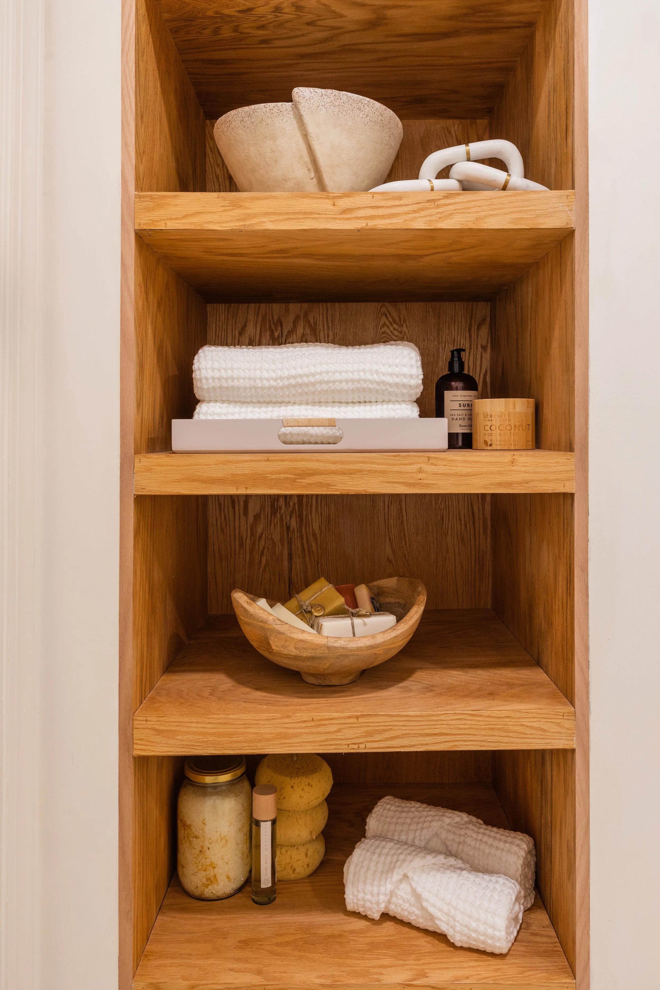 Wooden shelf with various bathroom accessories: top shelf has a beige bowl and a white chain, middle shelf has folded white towels, a lotion bottle, a soap bar, and a jar, bottom shelf has a jar, a spray bottle, and additional towels.