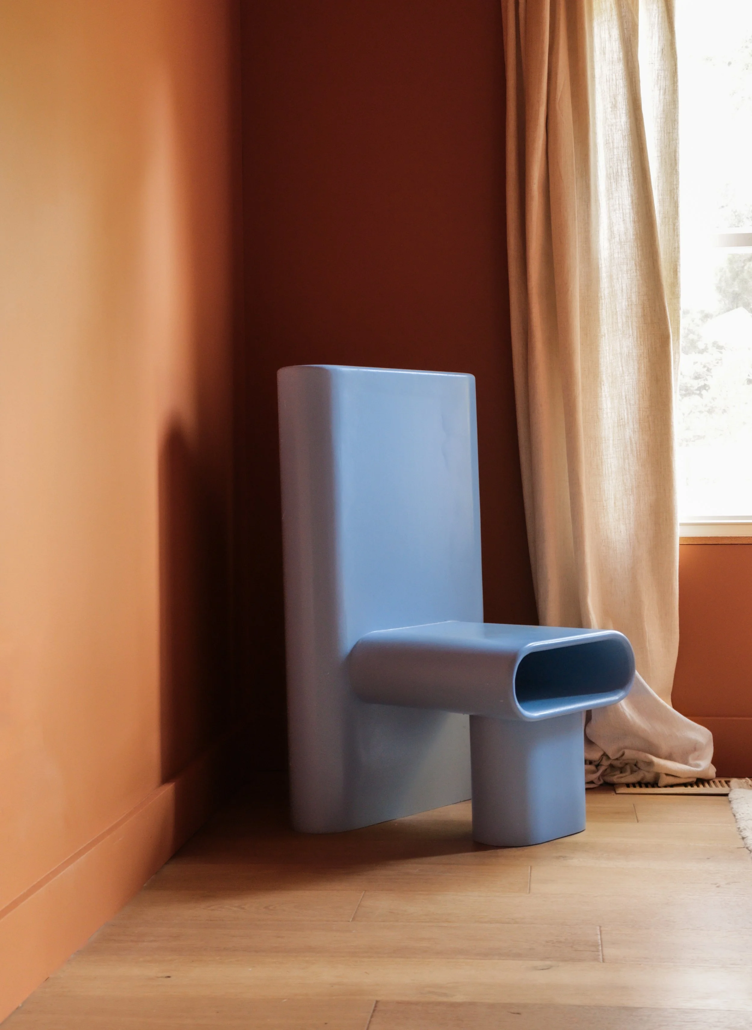 A blue sculptural hand made chair with a unique design, placed against a warm-colored orange wall near a window with beige curtains, in a cozy bedroom