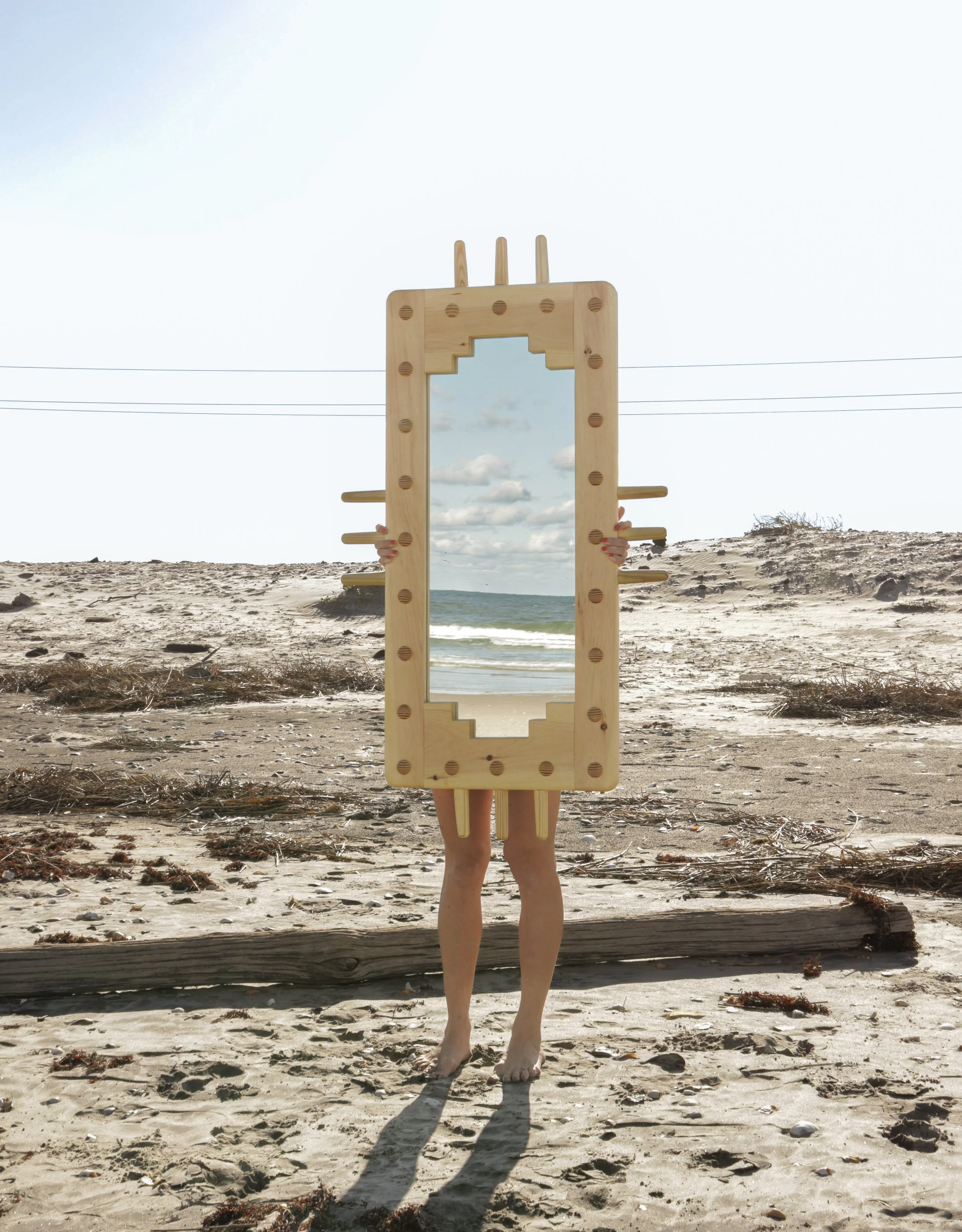 Person holding a mirror with a wooden frame in front of their body on a beach, with ocean and sky in the background.