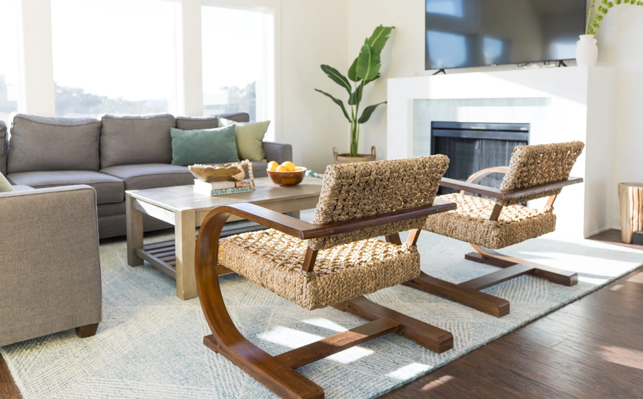 Living room with a gray couch, green pillows, a wooden coffee table with decorative objects and fruit, two woven armchairs, a large potted plant, a fireplace, a flat-screen TV, and a vase with greenery.