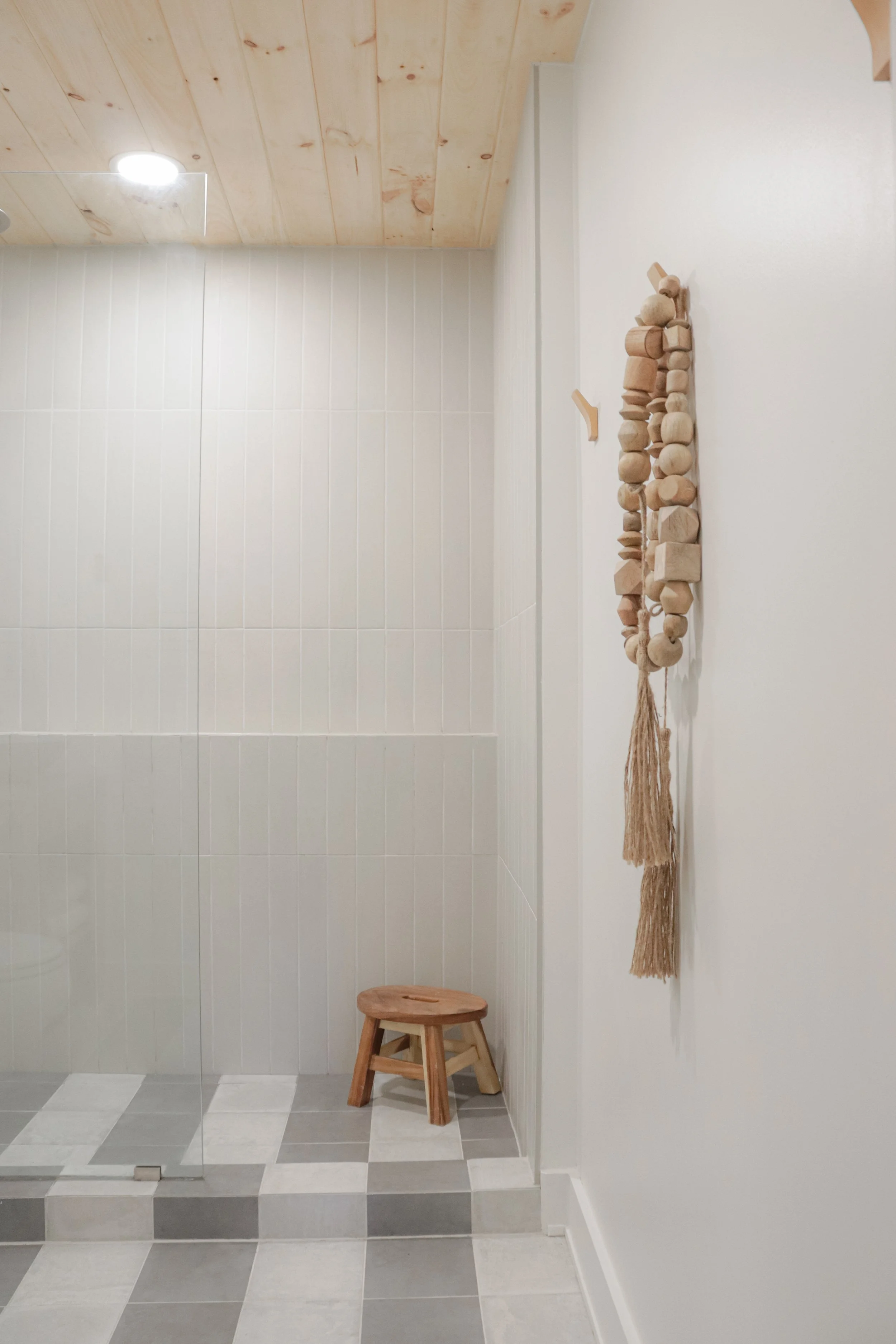 A small wooden stool in a bathroom with light-colored tiled walls and gray and white checkered floor tiles. There is a hanging decoration made of natural-colored wooden beads and shapes on the wall and a reflection of a light on a mirror or glass on 