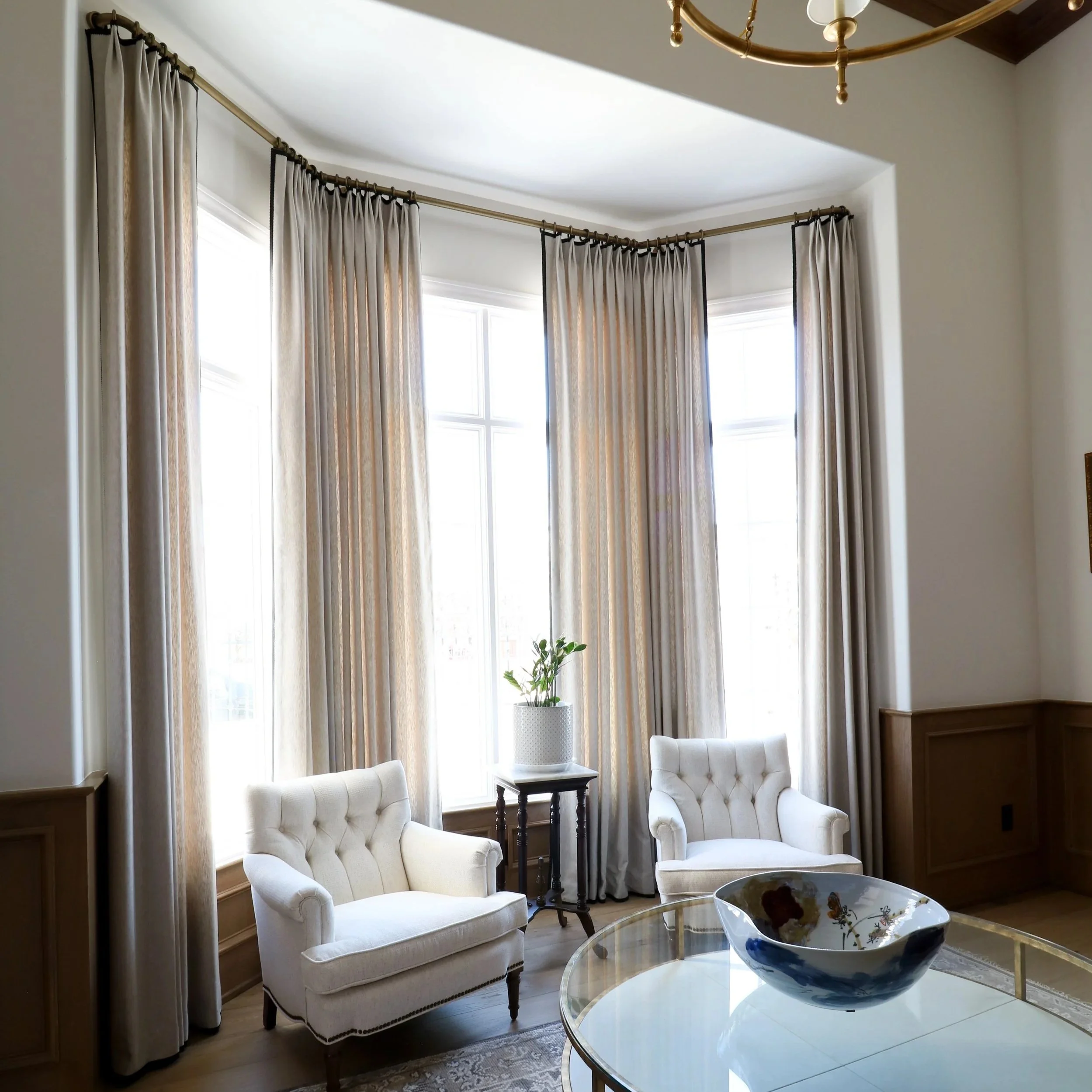 Bright living room with large windows framed by beige curtains, white tufted armchairs, a glass coffee table with a decorative bowl, a small side table with a potted plant, and a brass chandelier hanging from a vaulted ceiling.