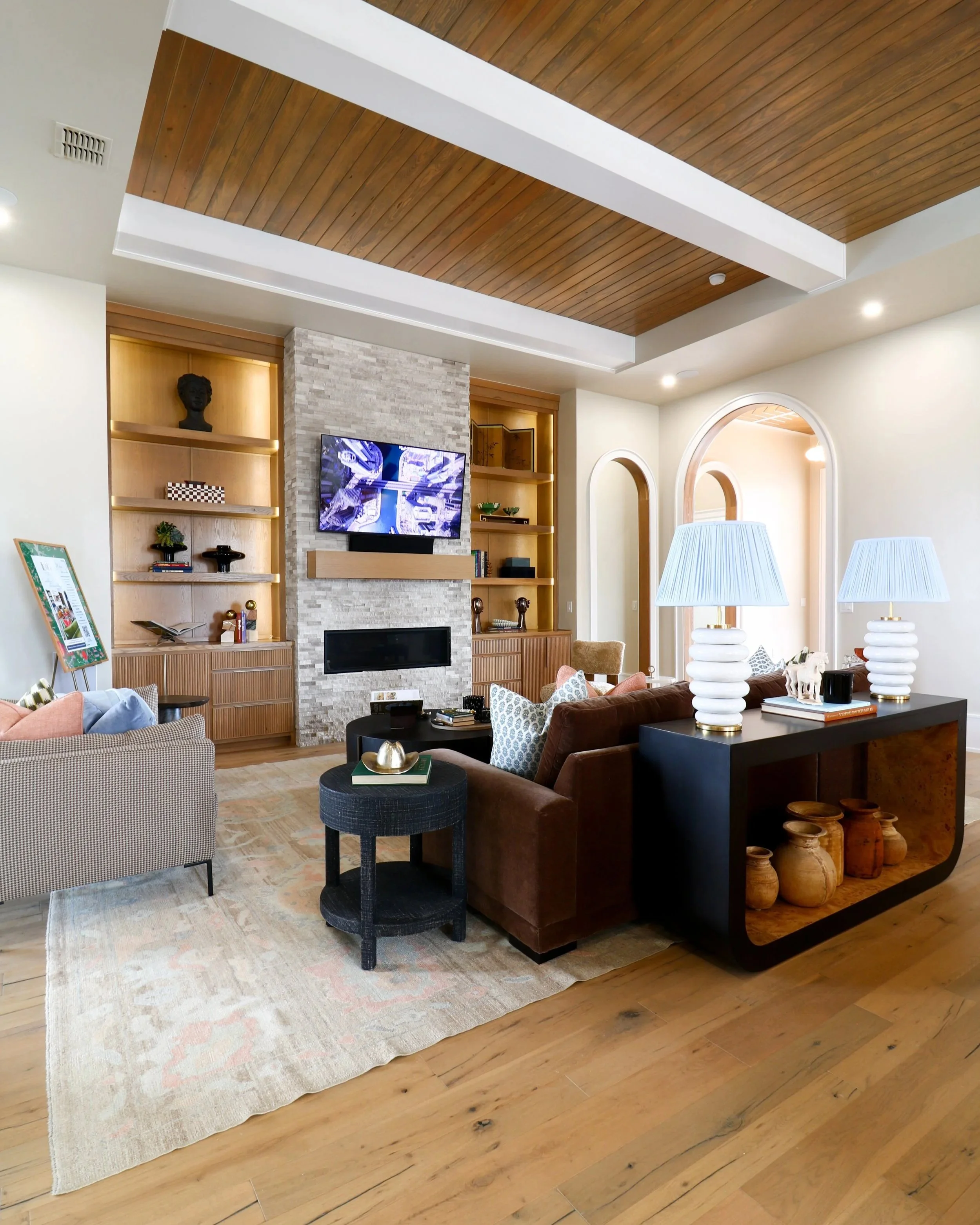 Living room with wood ceiling, built-in shelves, stone fireplace with television, armchair, sofa, side tables, lamps, and accessories.