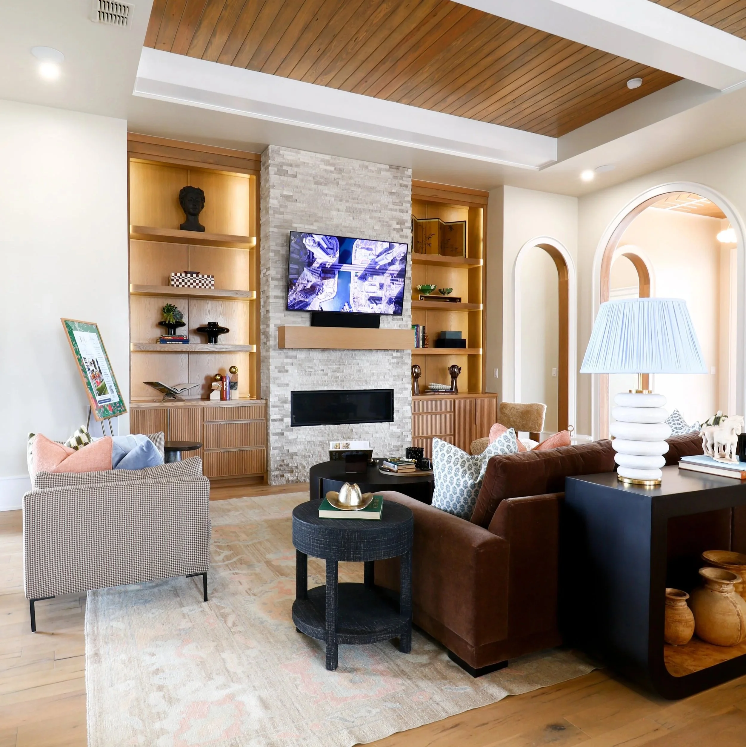A living room with a wooden ceiling, a flat-screen TV mounted on a stone fireplace, a bookshelf with decorative items, a beige armchair, a black side table with books and a gold object, and a brown sofa with patterned pillows.